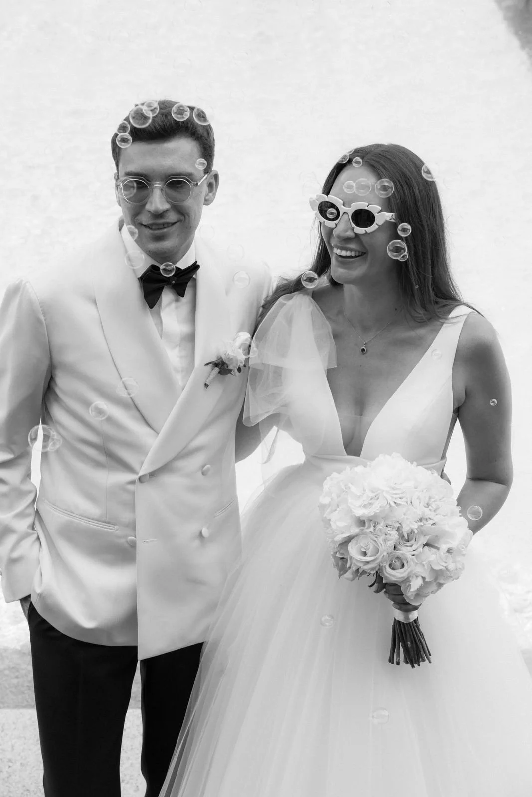 Ein glückliches Brautpaar trägt moderne Brillen und steht im Freien, während Seifenblasen um sie herum schweben, die Hochzeit humorvoll und fröhlich feiern.