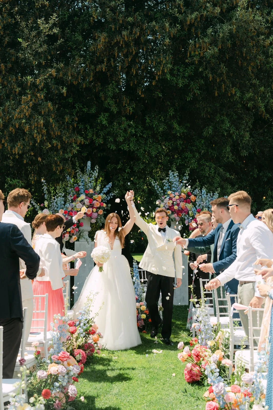 Hochzeit im Freien mit Braut und Bräutigam, umgeben von feiernden Gästen, Blumen und Dekoration, unter einem Baum.