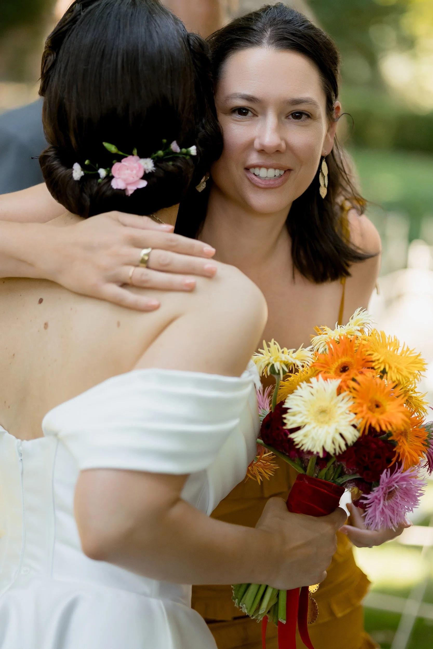 Eine Frau in einem weißen Kleid wird umarmt, während sie Blumen hält, im Hintergrund sind andere Personen und Natur. Es sieht nach einer Feier aus.