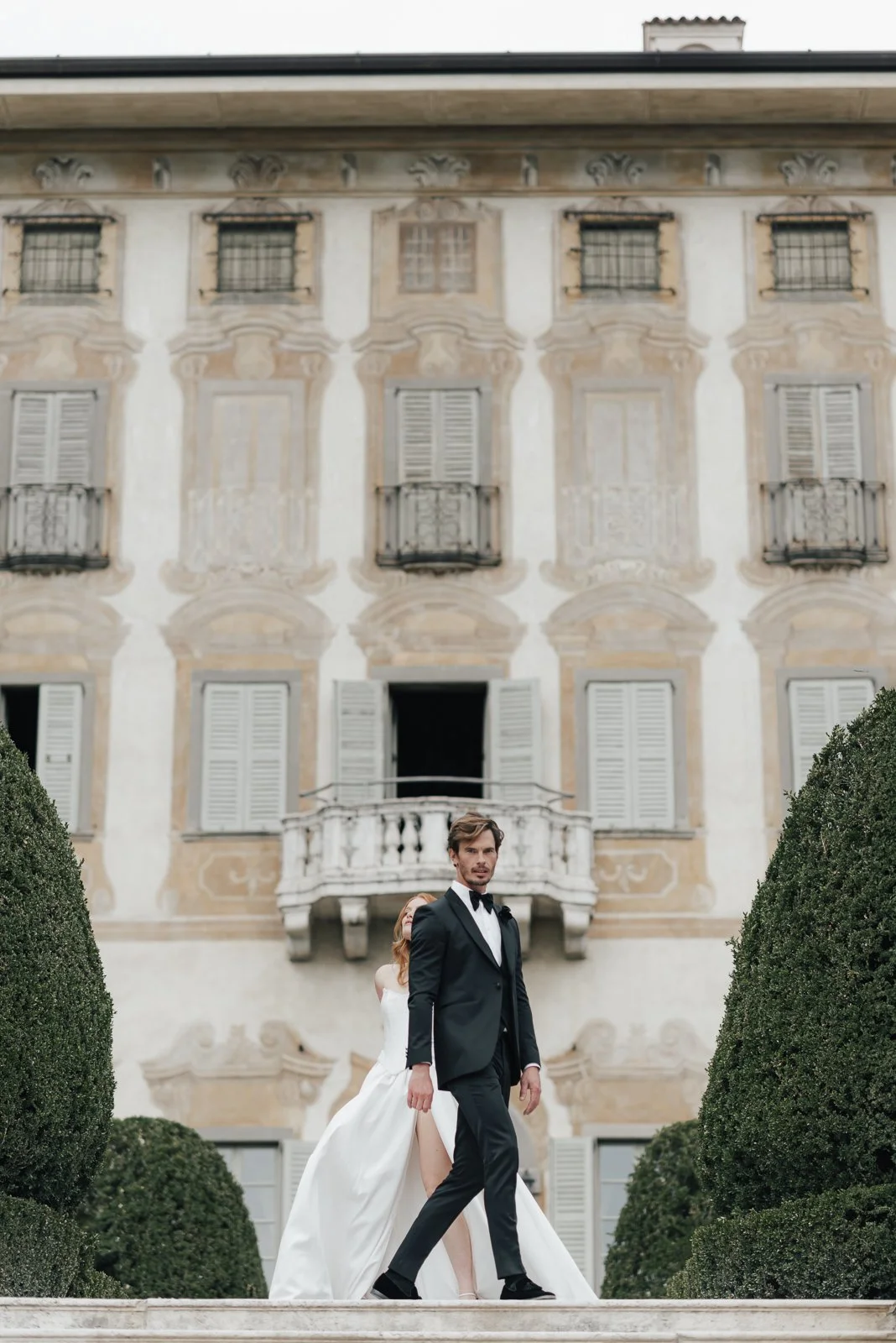 Ein gut gekleideter Mann in einem schwarzen Anzug und eine Frau in einem weißen Hochzeitskleid gehen auf einer Treppe vor einem historischen Gebäude mit Balkonen und Fensterläden. Es ist ein elegantes Hochzeitsfoto.