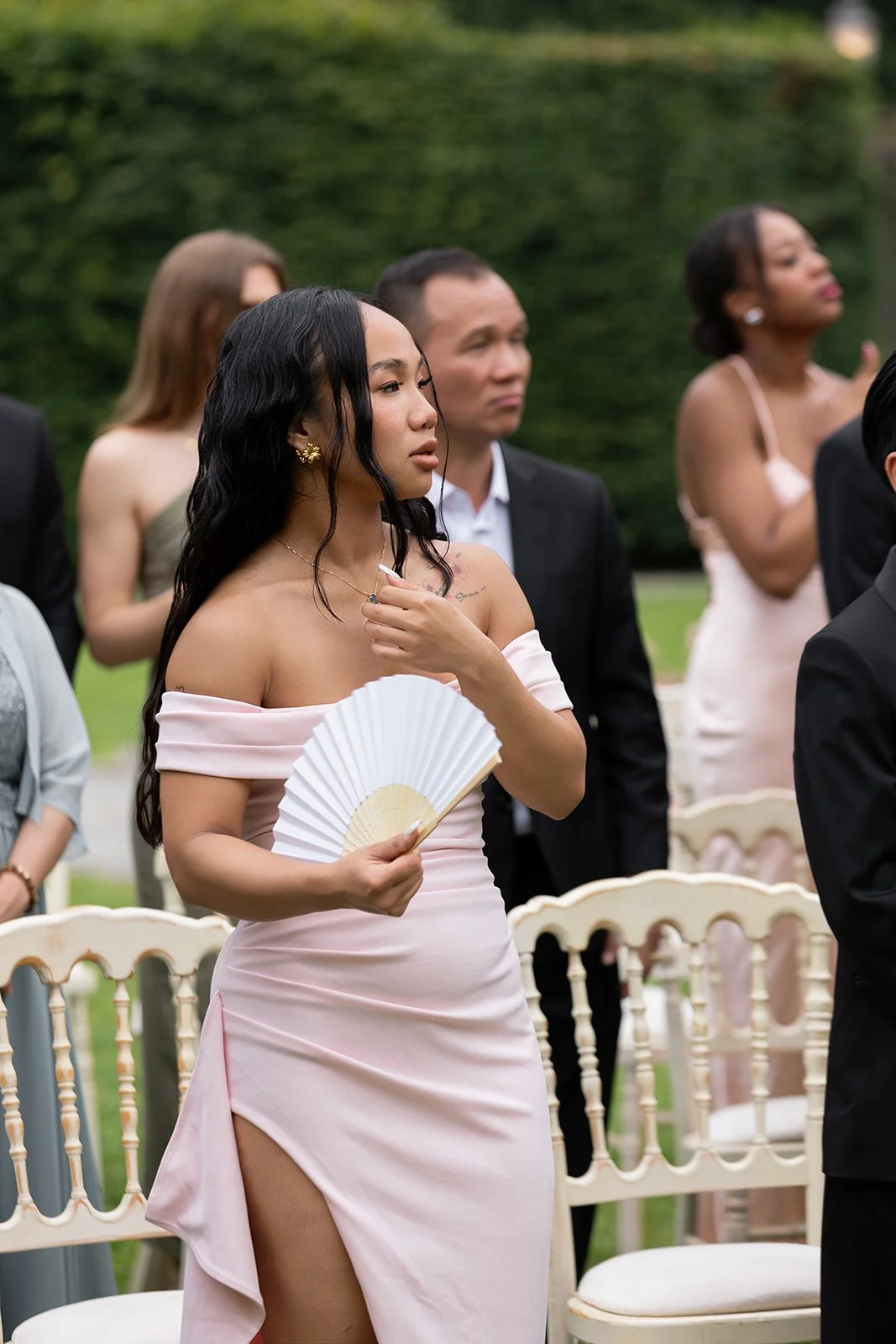 Junge Frau mit dunklen, lockigen Haaren trägt ein elegantes, off-shoulder, rosafarbenes Kleid mit hohem Schlitz und hält ein Handfächer bei einer formellen Veranstaltung im Freien.