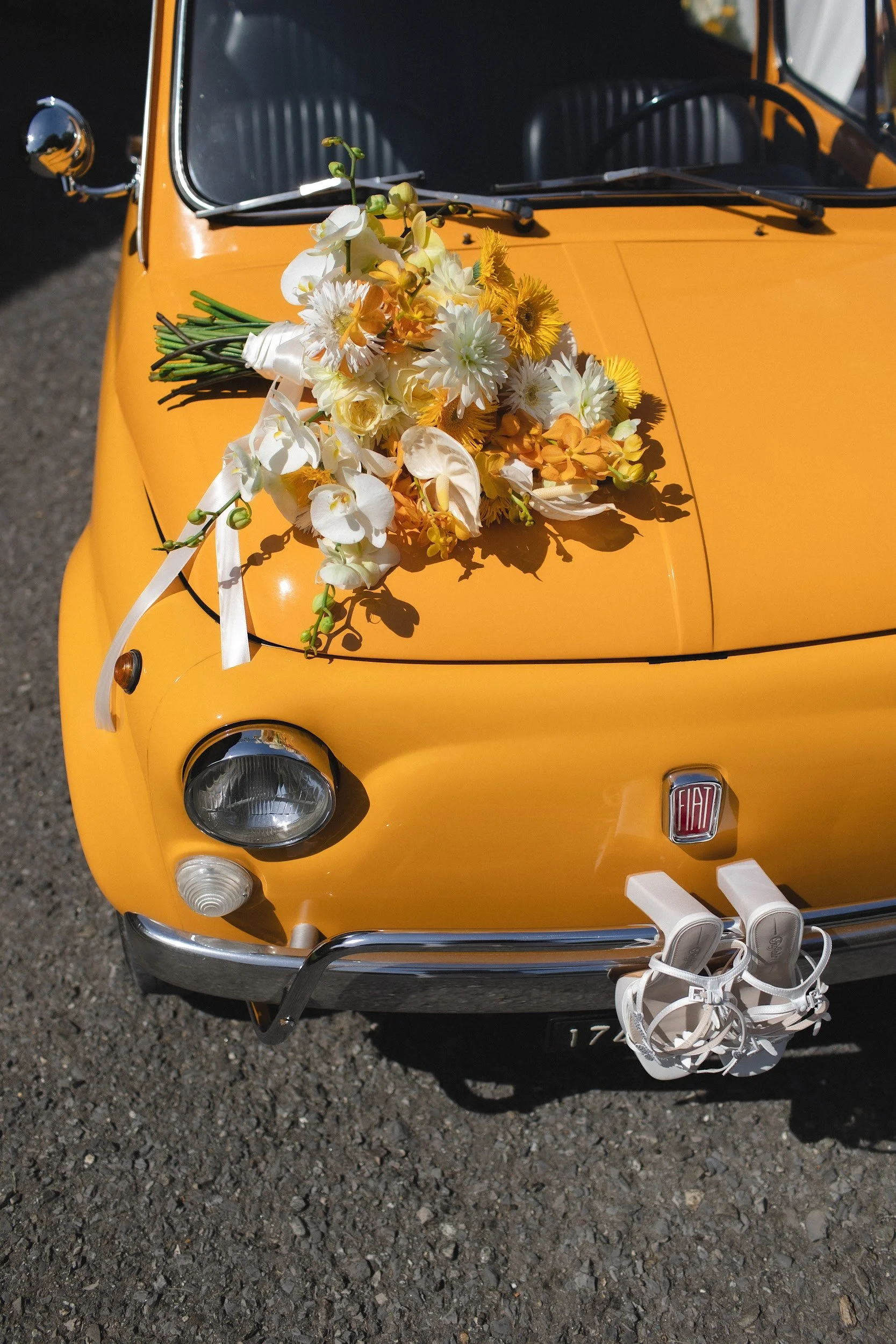 Gelbe Fiat-Auto mit Blumenarrangement auf der Motorhaube, weiße Schuhe hängen an der Stoßstange, im Hintergrund ein unbefestigter Boden.