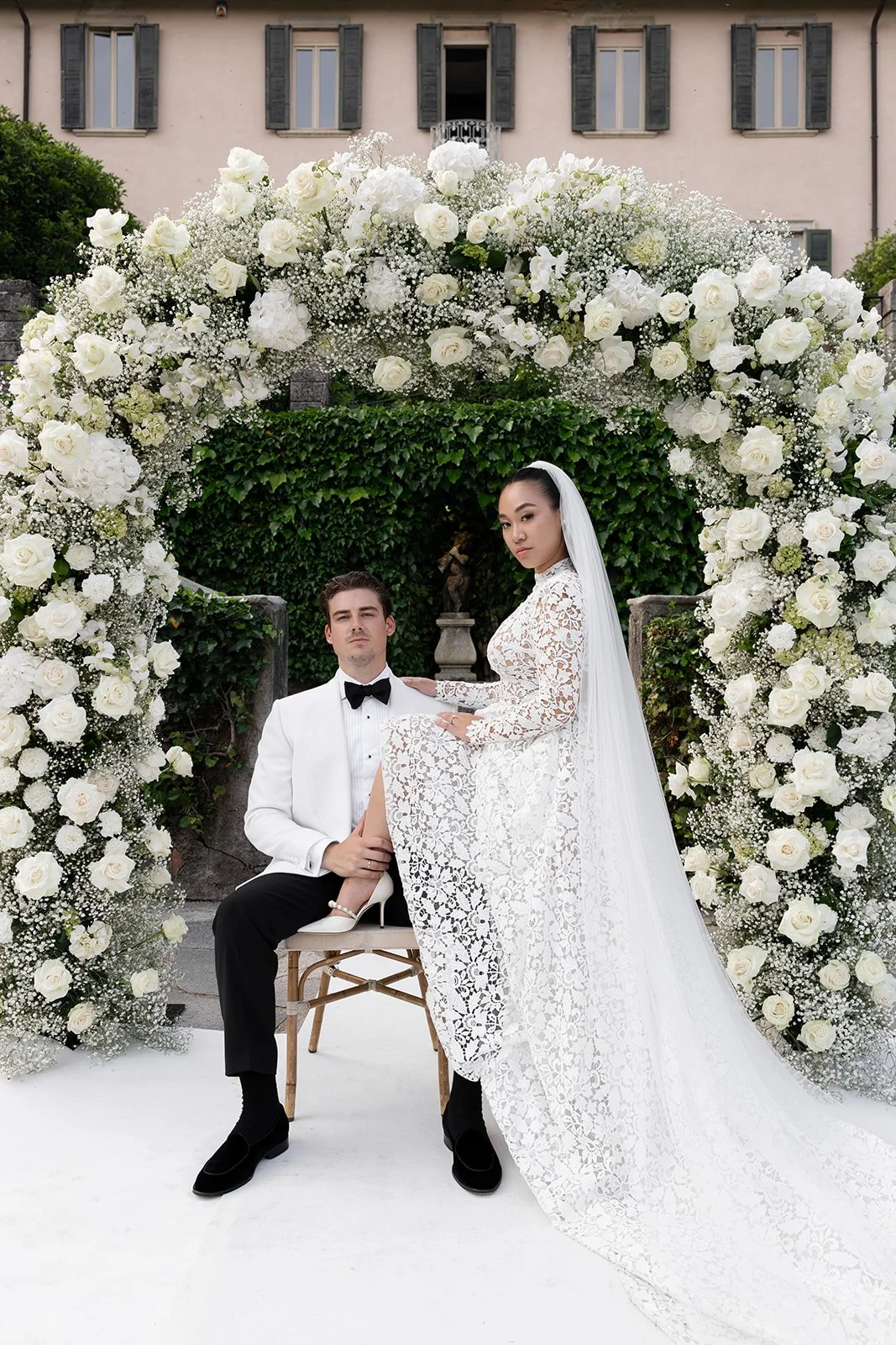 Ein heiratendes Paar vor einem Garten mit Blumenbogen, der aus weißen Rosen und anderen weißen Blumen besteht.