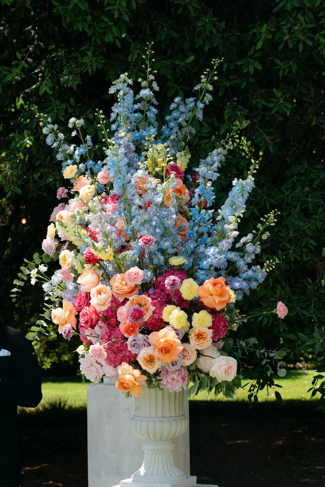 Bunter Blumenstrauß in einer weißen Vase vor einem dunklen grünen Busch.