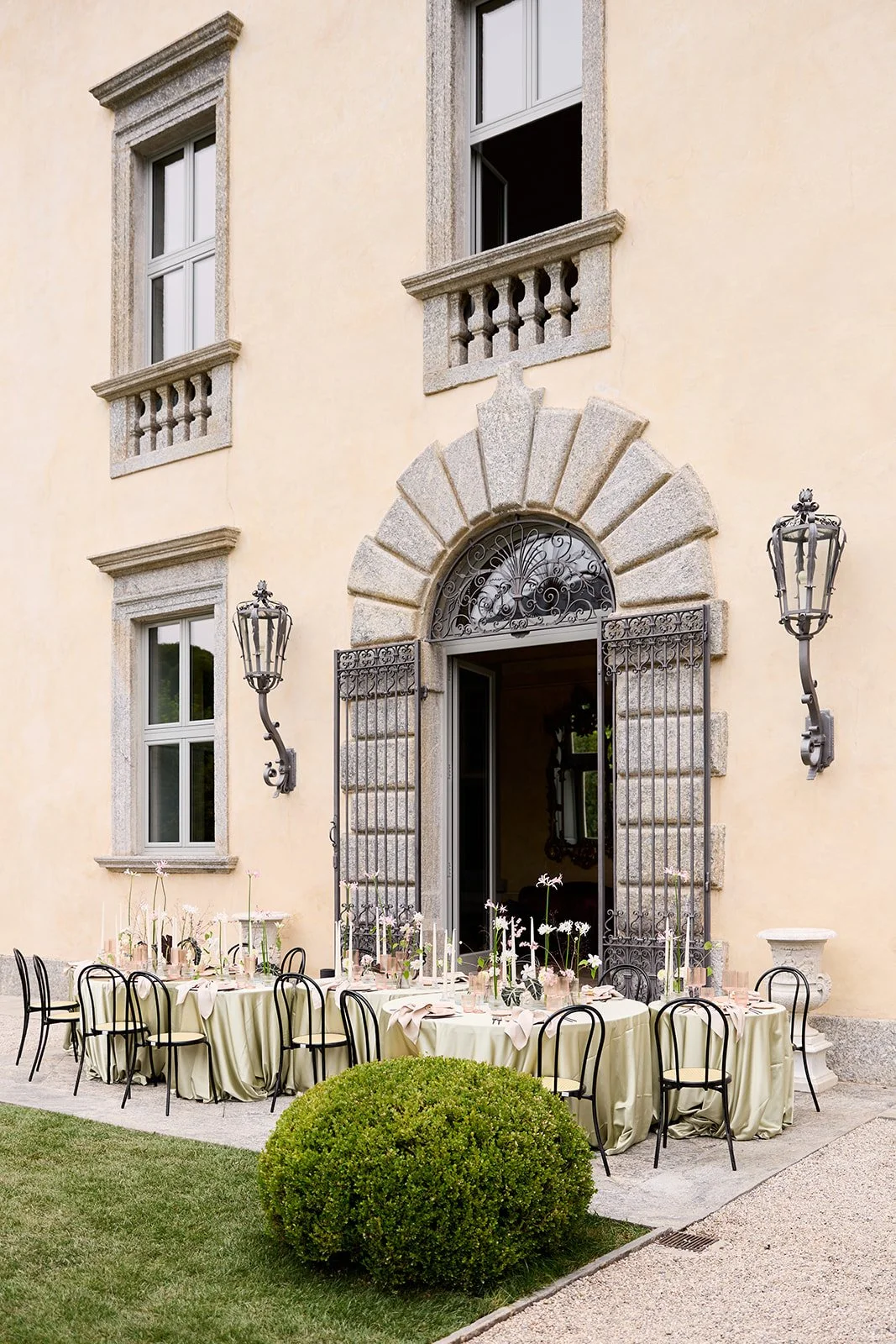 Eleganter Eingang eines Hauses mit kunstvoller Schmiedearbeit im Tor und an den Laternen, festlich gedeckter Sitzbereich mit runden Tischen und Stühlen im Außenbereich, umgeben von gepflegtem Grün.
