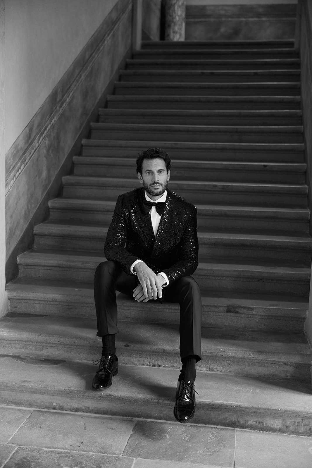 Ein Mann in einem schwarzen Pailletten-Jacket, flache schwarz Schuhe und Fliege sitzt auf einer Treppe, schwarz-weiß Fotografie.