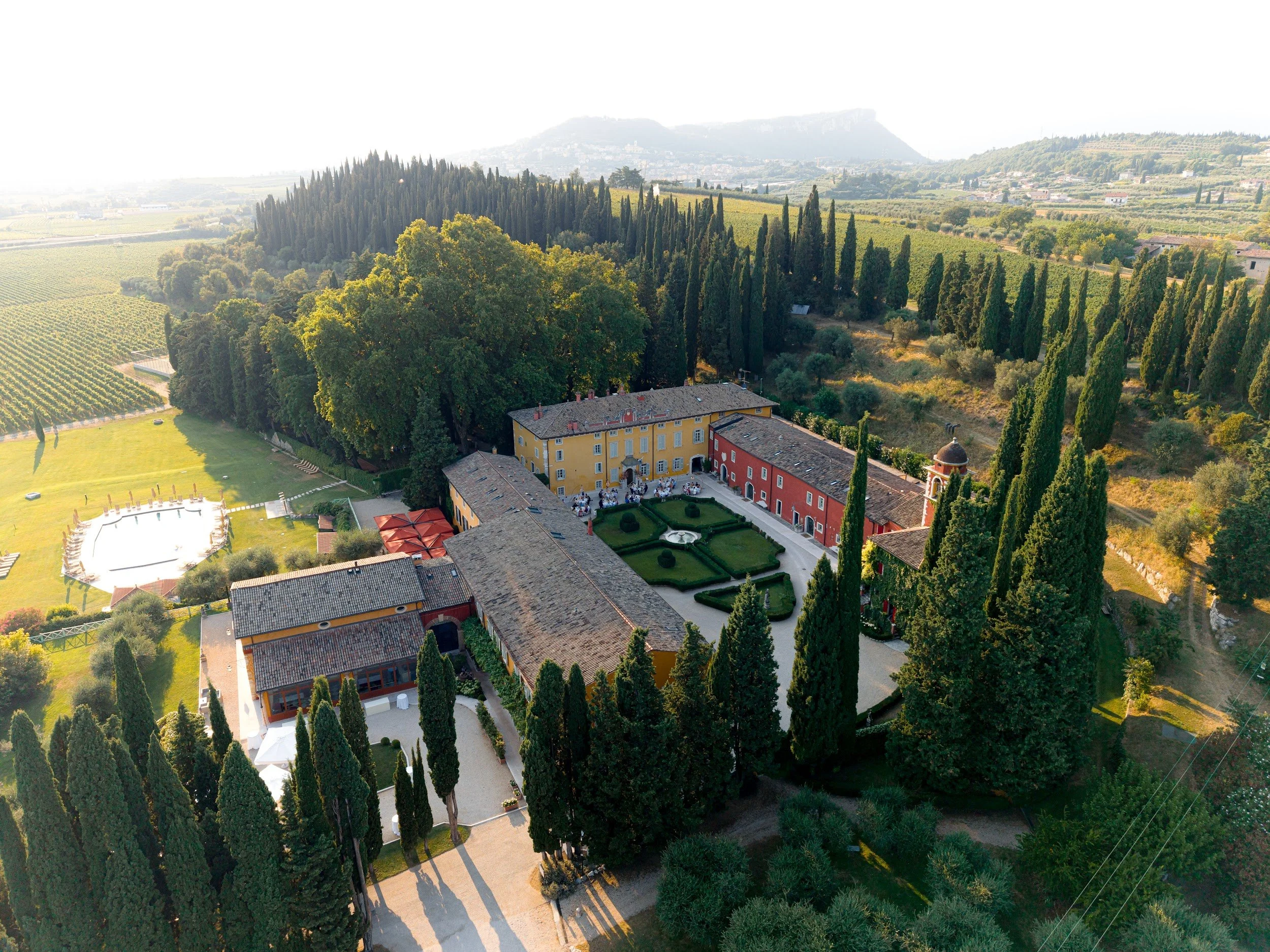 Blick auf eine ländliche Landschaft mit einem großen Garten, einer Villa und vielen hohen Bäumen, umgeben von Weinbergen und Hügeln im Hintergrund.