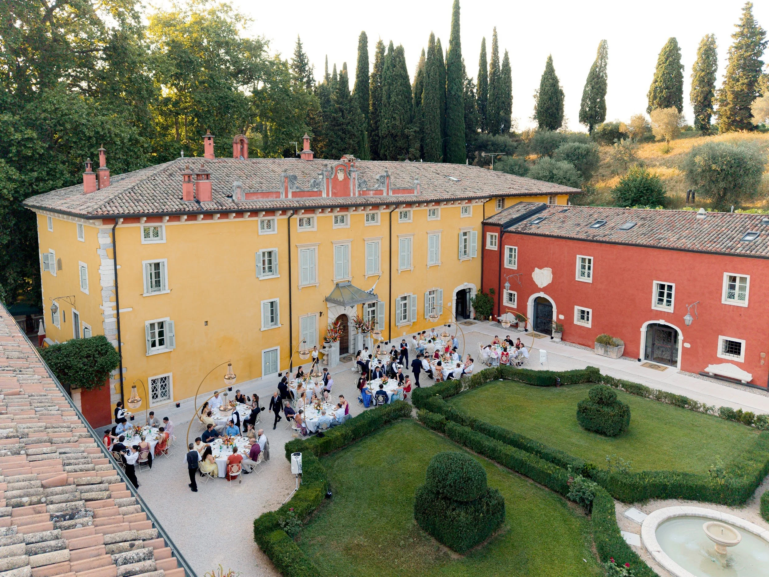 Eine Terrasse im Freien mit mehreren Tischen, an denen Menschen bei einem festlichen Essen sitzen. Im Hintergrund eine farbige Gebäude-Front mit roten und gelben Wänden. Im oberen Bereich des Bildes sind hohe Kiefern und andere Bäume zu sehen.