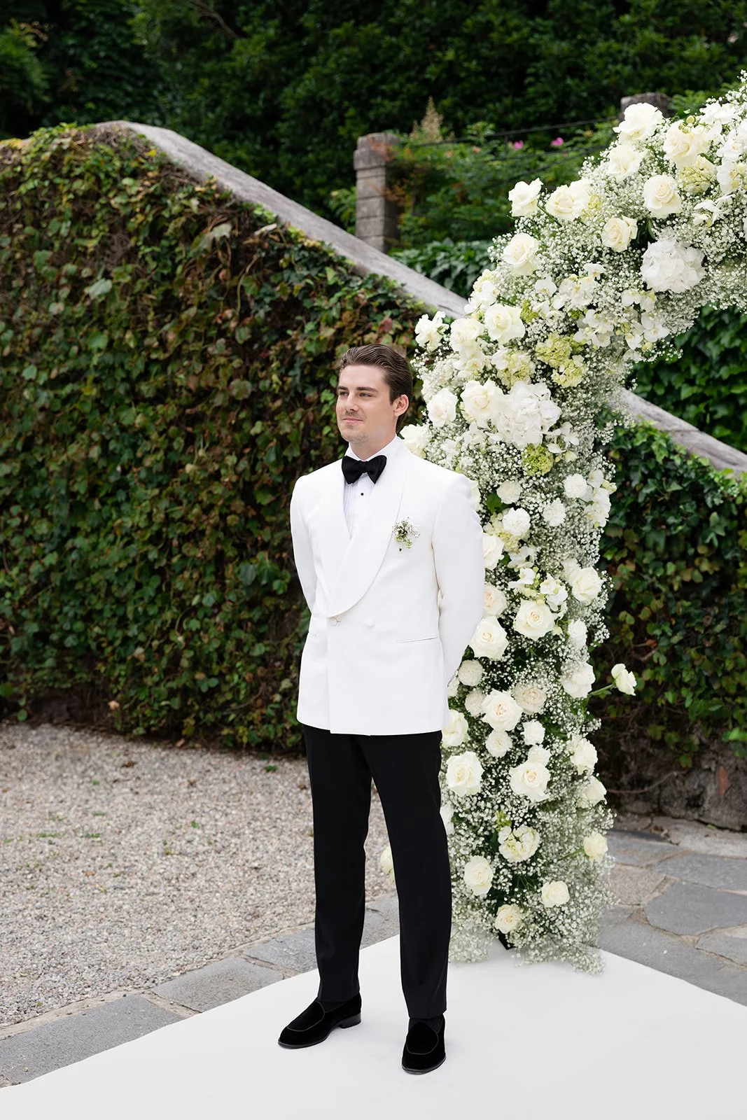 Junger Mann in weißem Anzug mit schwarzer Fliege bei einer Trauung, steht vor einem großen Blumenbogen aus weißen Rosen und Babys breath, im Garten. 