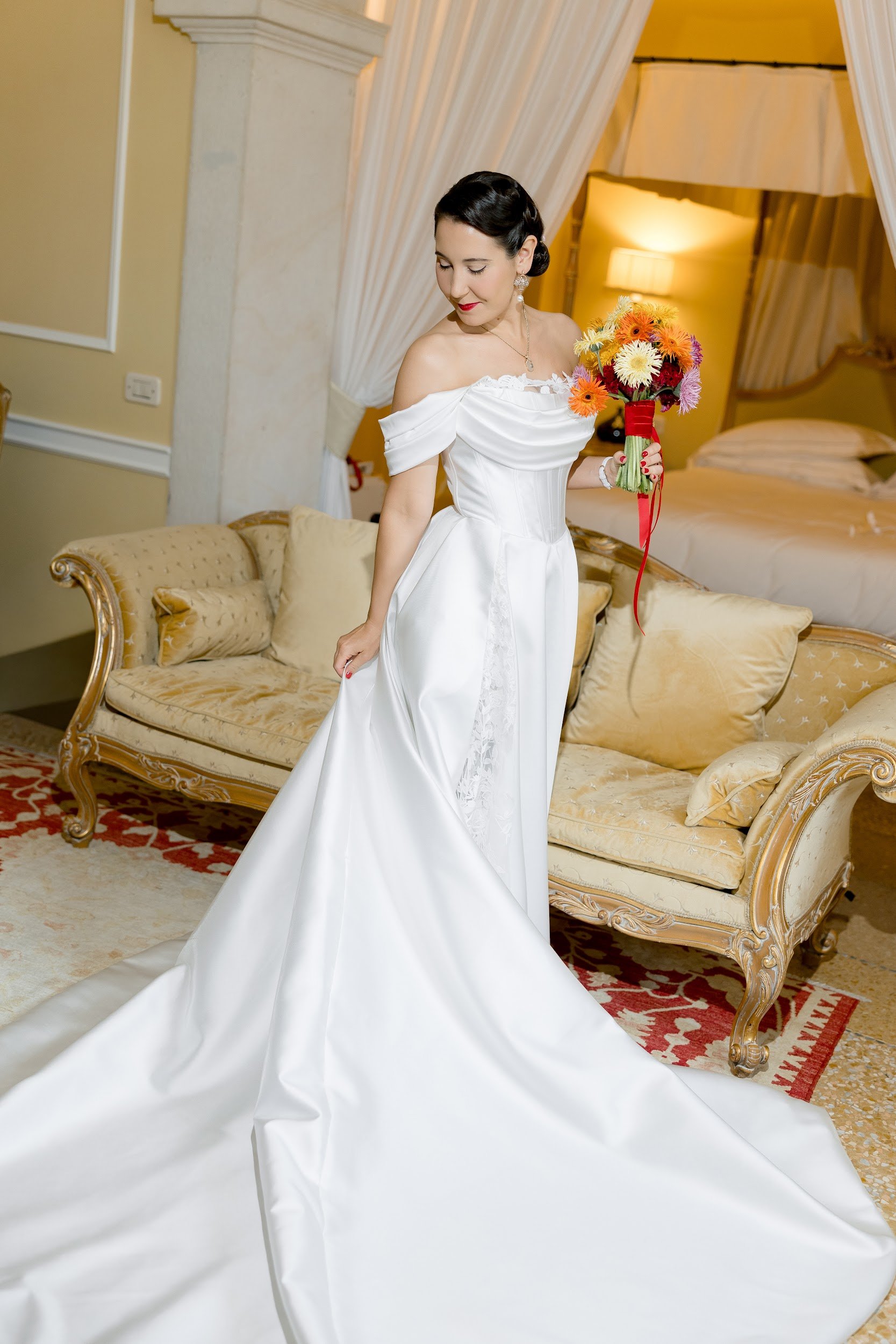 Eine Frau in einem weißen Brautkleid hält einen bunten Blumenstrauß und steht in einem gemütlichen, elegant eingerichteten Zimmer.