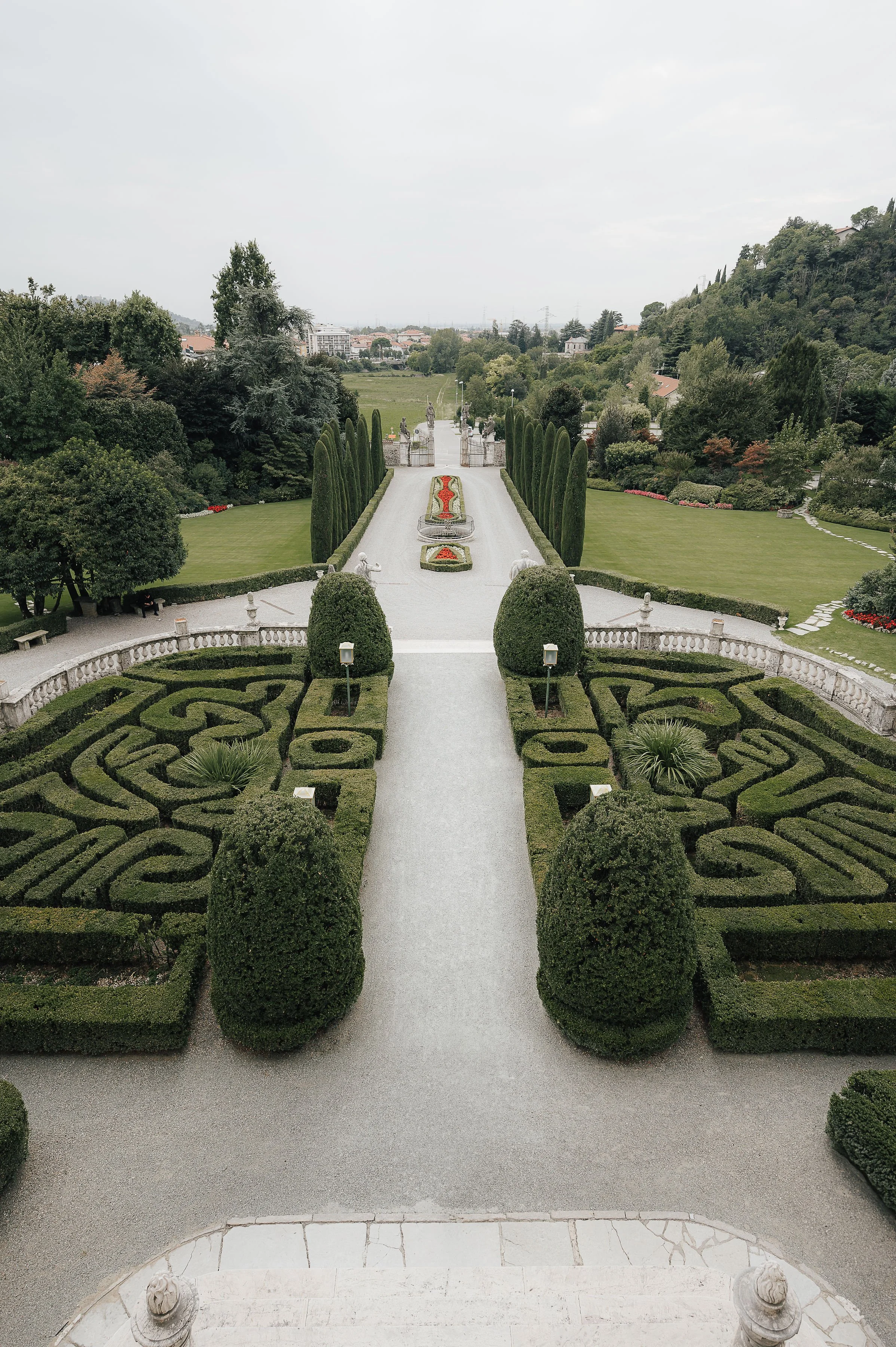 Gartenanlage mit einem Wasserbecken, formgeschnittenen Hecken und Bäumen, Statuen und einer Treppe im Vordergrund, im Hintergrund grüne Hügellandschaft und Städte
