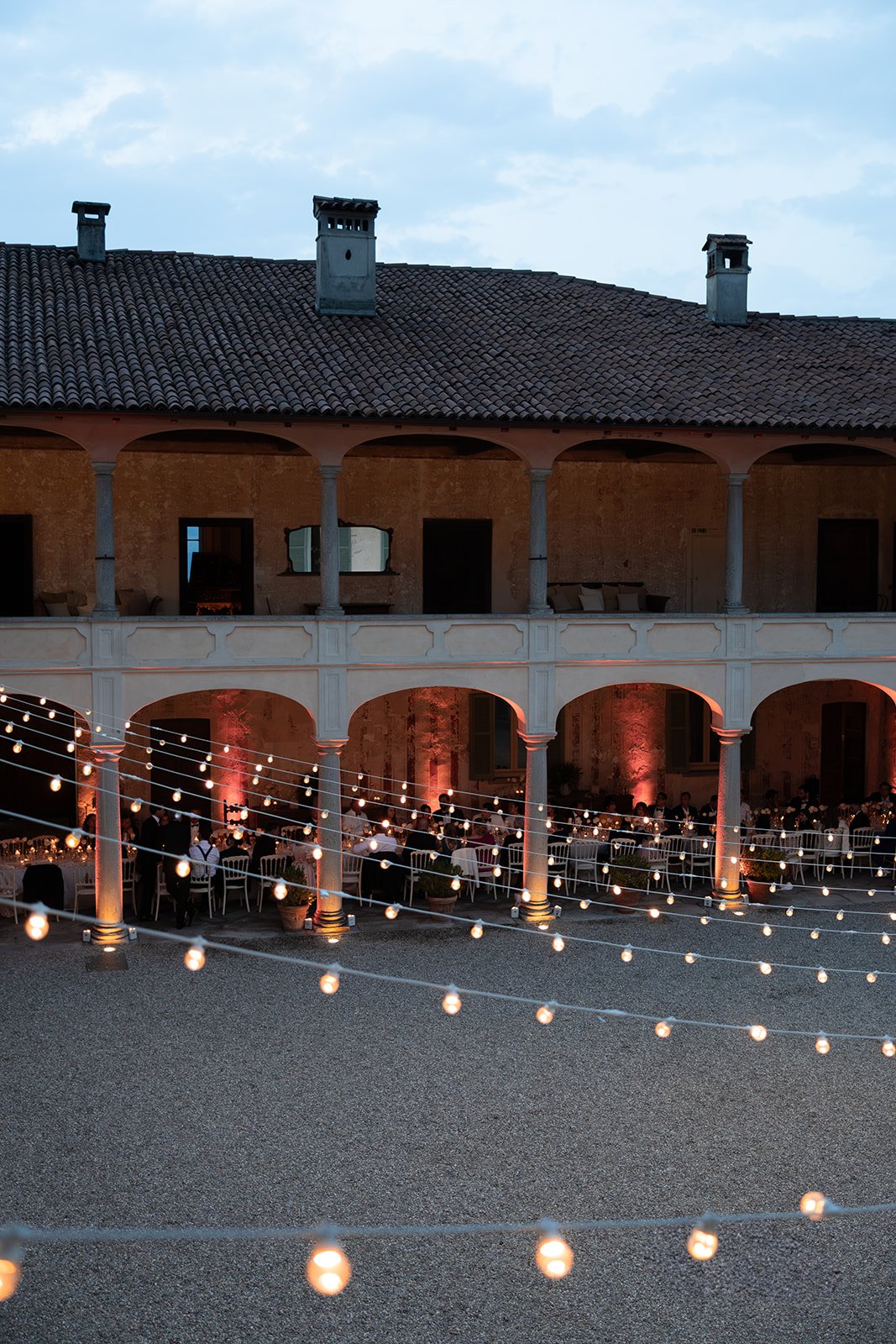 Ein festlich dekorierter Innenhof mit mehreren Tischen, Lichterketten und Gästen bei einer Abendveranstaltung in einem historischen Gebäude mit einem höheren Balkon im Hintergrund.