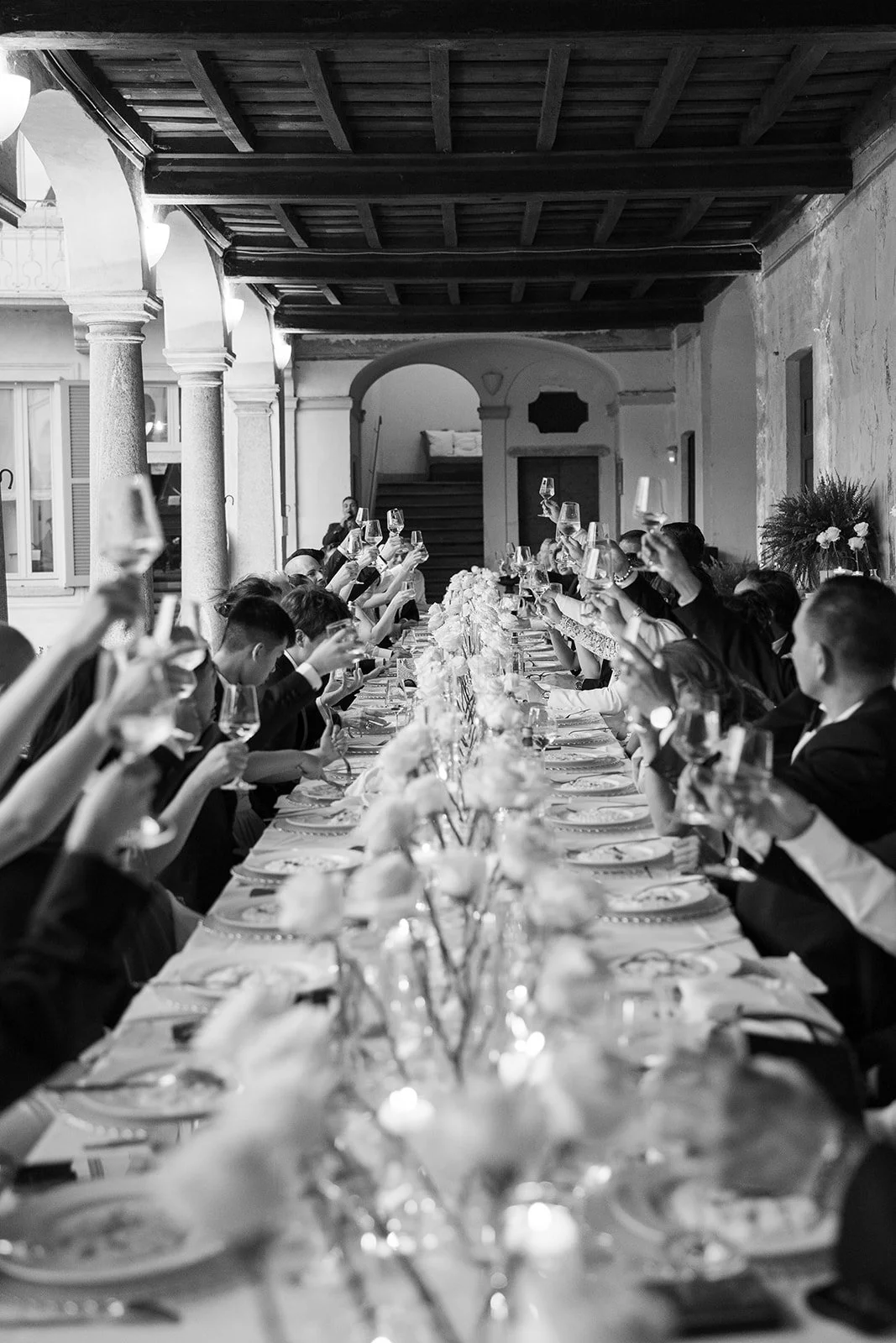 Eine schwarze und weiße Aufnahme einer festlichen Dinerszene mit vielen Gästen, die an einem langen Tisch sitzen und mit Weingläsern anstoßen.
