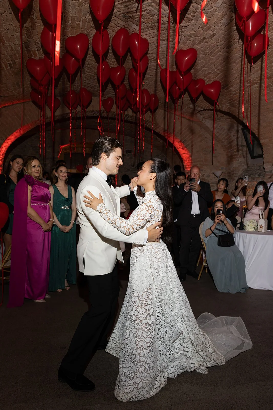 Ein Brautpaar tanzt bei einer Hochzeit. Im Hintergrund sind rote Herzballons aufgehängt und Gäste, die Fotos machen.