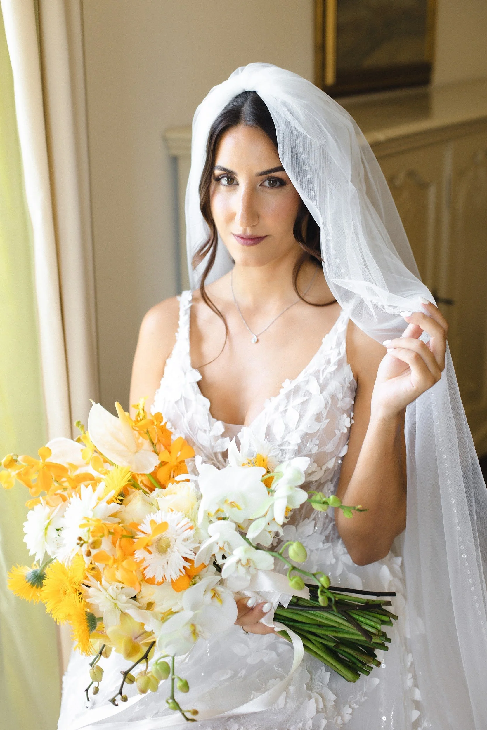 Eine junge Frau in einem weißen Brautkleid mit Blumenmuster hält einen großen Blumenstrauß mit weißen, gelben und orangefarbenen Blumen.