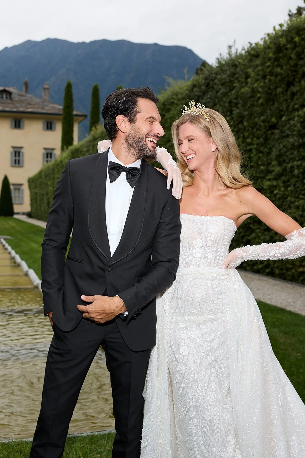 Ein lachendes Brautpaar bei einer Hochzeit im Freien, mit einem Mann im Smokingsjacke und einer Frau in einem weißen Hochzeitskleid, im Hintergrund ein Gebäude, Bäume und Berge.