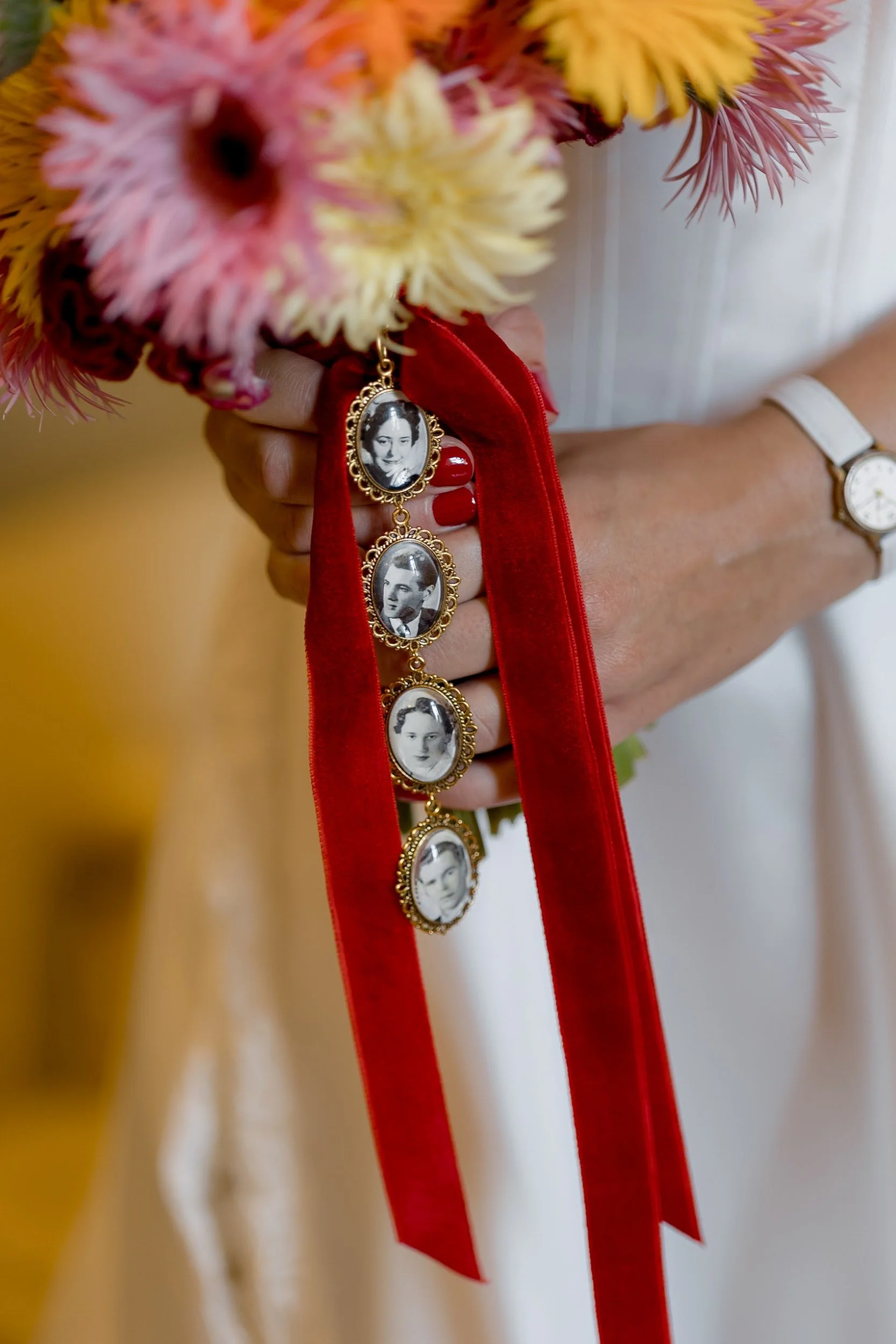 Eine Hand hält einen Blumenstrauß mit bunten Blumen und einer roten Schleife mit schwarz-weißen Fotoplaketten. Auf dem Handgelenk trägt die Person eine weiße Armbanduhr.