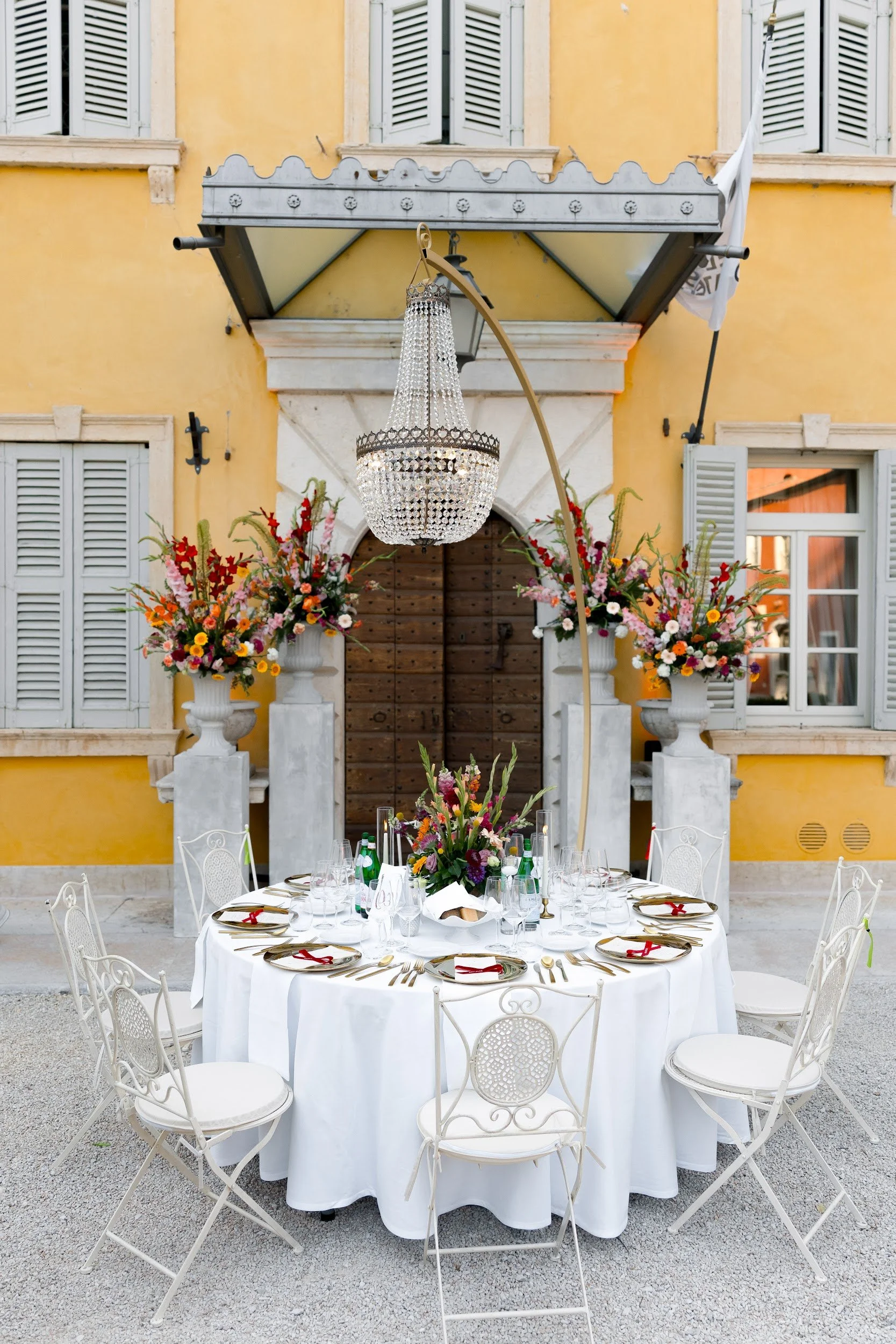 Eleganter Gala-Tisch im Freien mit weißen Tischdecken, goldenen Bestecken, Gläsern und roten Servietten, mit großen Blumenarrangements und einem Kronleuchter, vor einem gelben Gebäude mit weißen Fenstern und Türen.