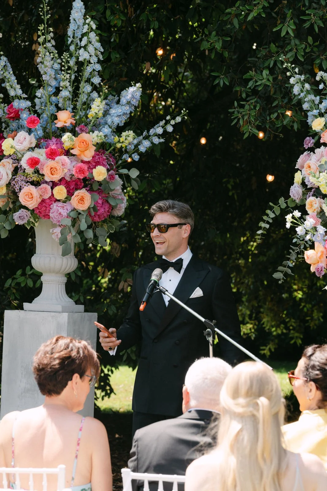 Ein Mann im schwarzen Anzug, Sonnenbrille, lächelnd, hält ein Mikrofon und eine Notizkarte während einer outdoor Rede bei einer Hochzeit. Im Hintergrund große Blumenarrangements mit Rosen, Hortensien und anderen Blumen, umrahmt von grünen Bäumen.