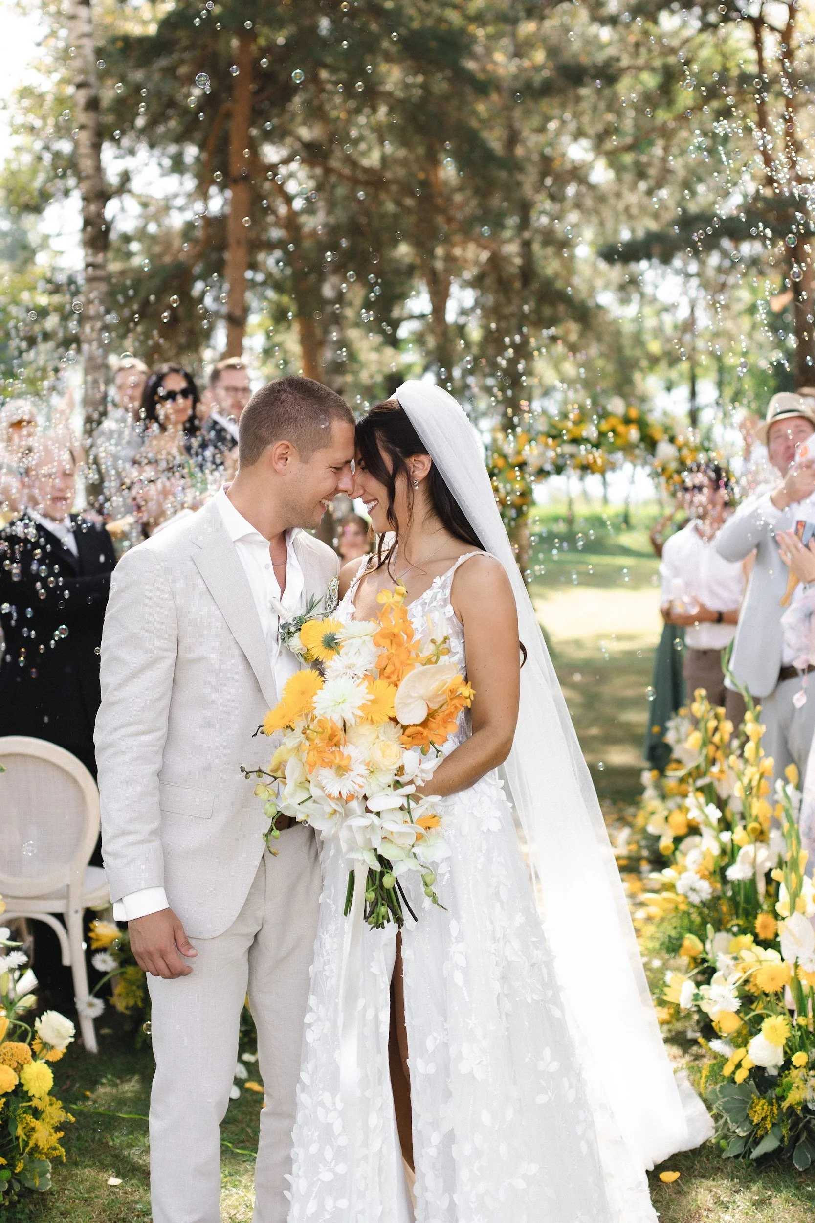 Ein frisch verheiratetes Paar bei einer Gartenhochzeit, umgeben von Freunden und Familie, sich an der Nase berührend, mit vielen gelben und weißen Blumen im Hintergrund.