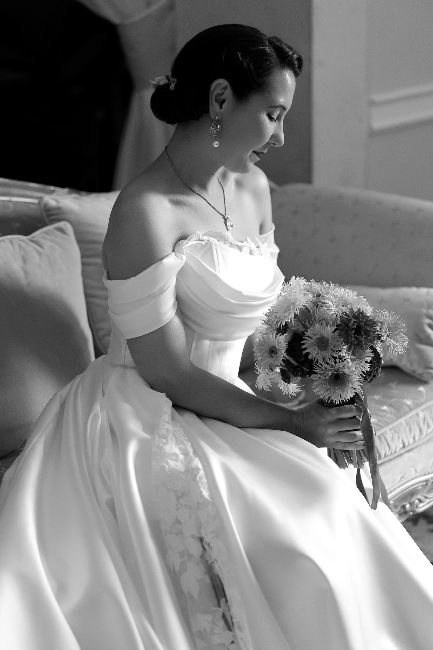 Schwarz-weiß-Foto einer glücklichen Frau in einem Hochzeitskleid, die auf einem Sofa sitzt und eine kleine Blumenstrauß hält.