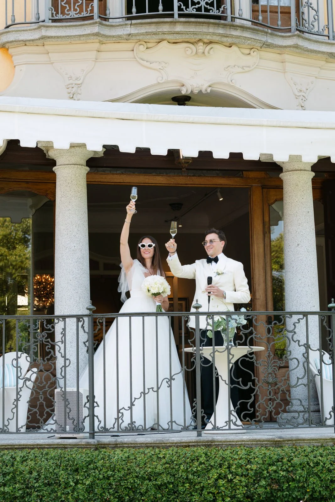 Ein frisch verheiratetes Paar auf einem Balkon, beide in formeller Hochzeitskleidung, stoßen mit Sektgläsern an, die Braut hält einen Blumenstrauß.