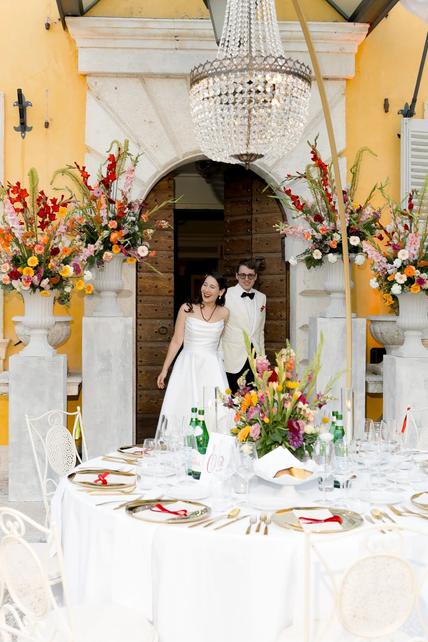 Ein Brautpaar, ein Mann in Smoking und eine Frau in Brautkleid, stehen vor einer Tür mit Holzplatte, flankiert von großen Blumenvasen mit bunten Blumen. Im Vordergrund ein festlich gedeckter Tisch mit weißen Tischdecken, Blumenstrauß, Wein, Wasser und Besteck.