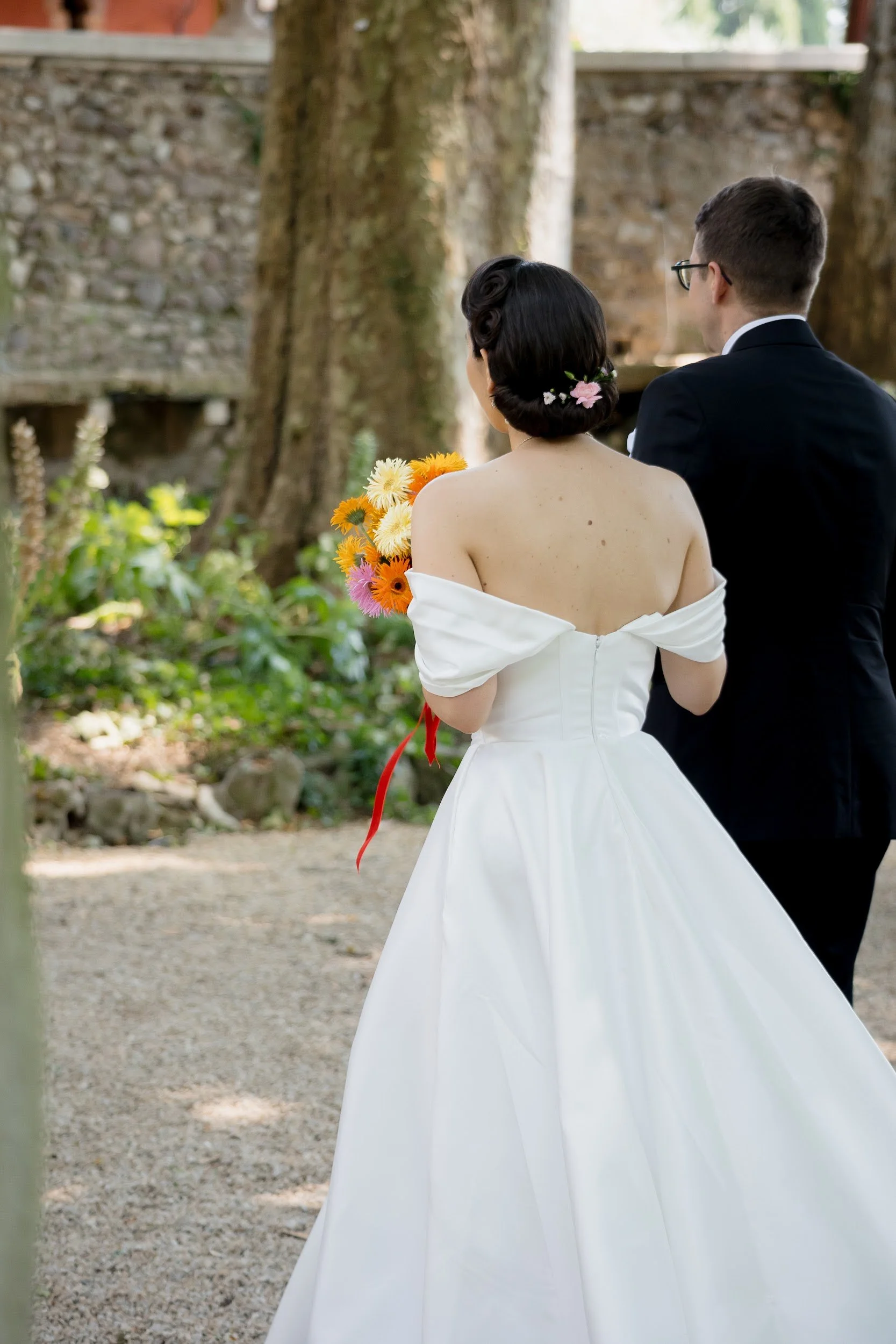 Ein Brautpaar steht im Freien vor einer Baumreihe. Die Braut trägt ein weißes Brautkleid mit off-shoulder-Design und hat einen Blumenstrauß in der Hand. Der Bräutigam trägt einen schwarzen Anzug und Brille. Die Szene ist im natürlichen Licht aufgenom