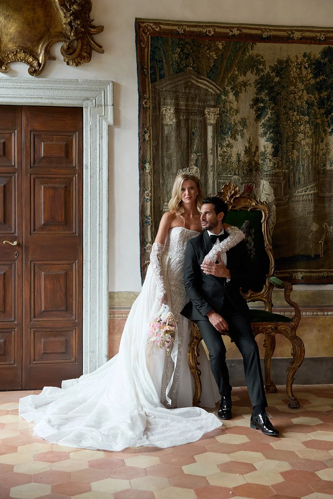 Hochzeitsfoto eines Brautpaares in eleganter Kleidung, sitzend und stehend vor einem historischen, kunstvollen Hintergrund.