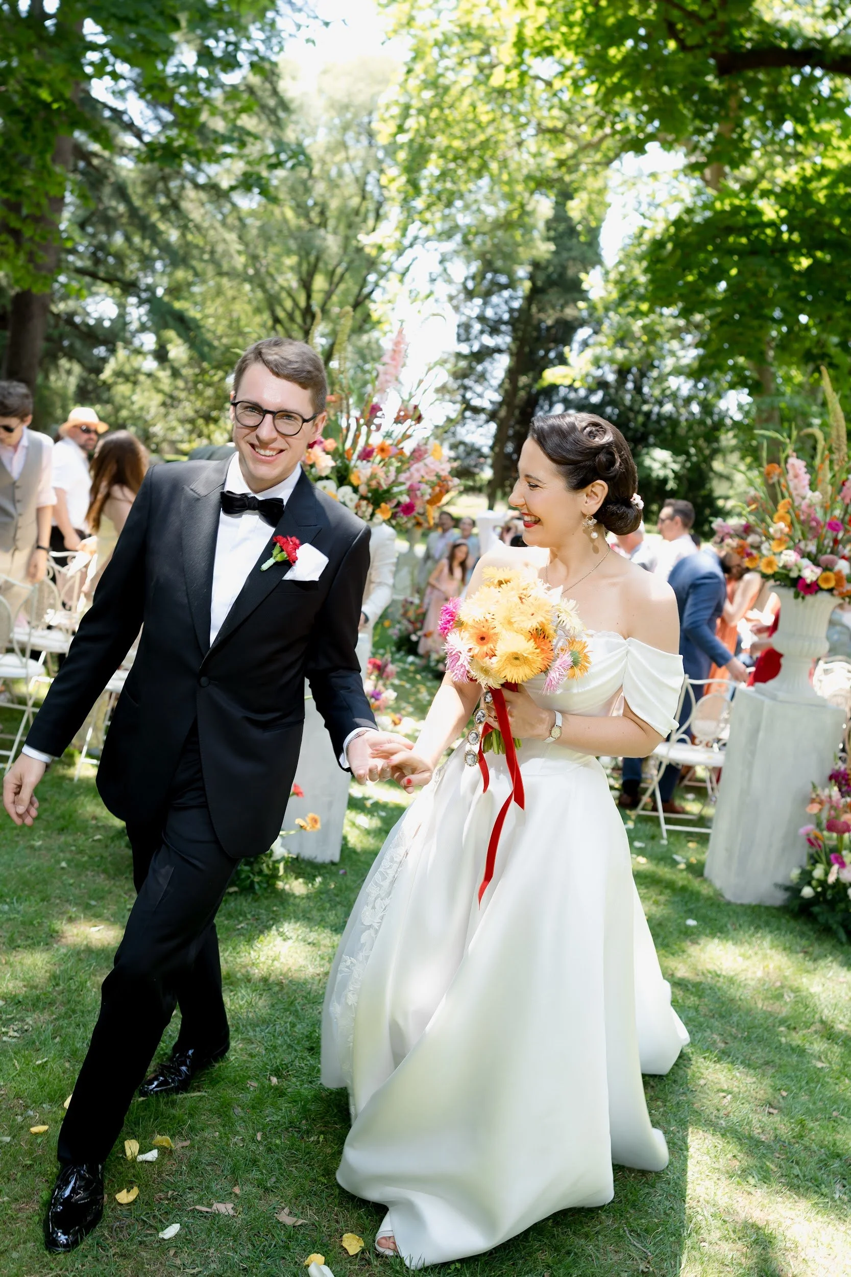 Ein Brautpaar läuft bei einer Hochzeit im Freien durch den Garten, umgeben von Gästen und bunten Blumen. Der Bräutigam trägt einen schwarzen Anzug mit Fliege, die Braut trägt ein langes weißes Kleid und hält einen Blumenstrauß.