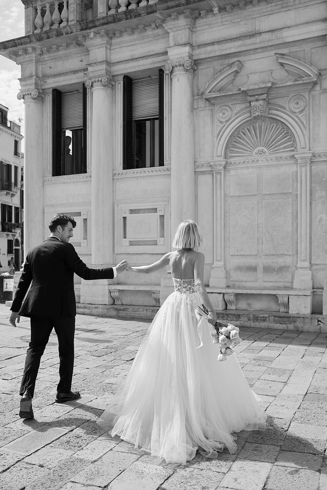 Ein Mann und eine Frau in einem Brautkleid laufen auf einer gepflasterten Straße vor einem historischen Gebäude mit Säulen. Die Frau hält einen Blumenstrauß in der Hand.