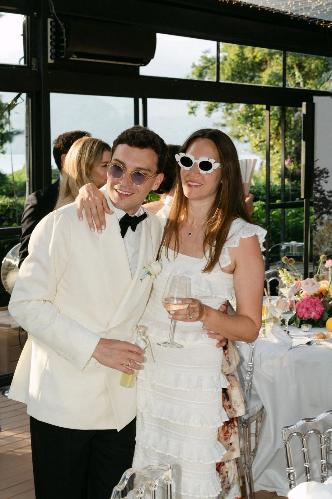 Ein Mann und eine Frau bei einer Feier, beide tragen weiße Kleidung und Accessoires im Retro-Stil, die Frau hält ein Weinglas. Sie stehen nebeneinander und lächeln. Im Hintergrund sind Dekorationen und andere Gäste sichtbar.