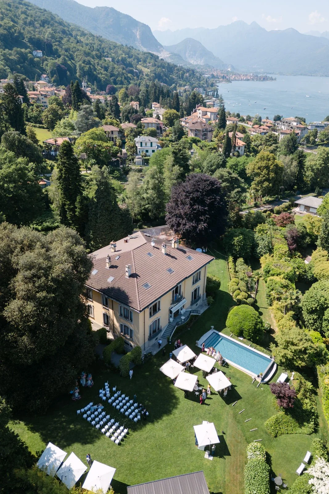 Ein historisches Gebäude mit Eingang, umgeben von einem Garten mit sitzgelegenheiten, einem Pool und einer Hängematte, in einer bewaldeten Gegend an einem See mit Bergen im Hintergrund.