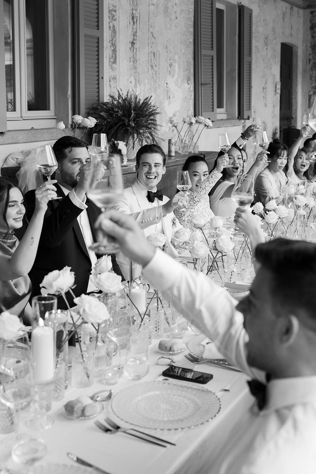Menschen bei einer Hochzeitsfeier, die mit Weingläsern anstoßen, im Hintergrund ein Tisch mit Blumen und Dekorationen.