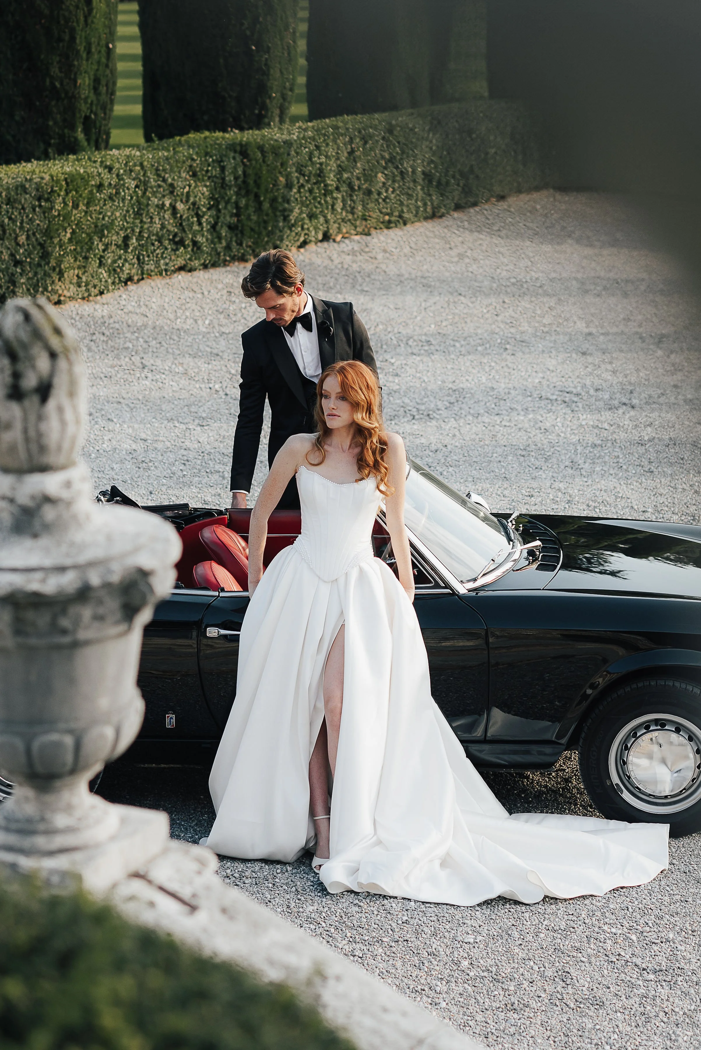 Braut in weißem Hochzeitskleid posiert vor einem schwarzen Oldtimer, während ein Mann im schwarzen Anzug und Fliege im Hintergrund steht. Der Hintergrund zeigt eine gepflegte Gartenlandschaft mit Hecken und Bäumen.