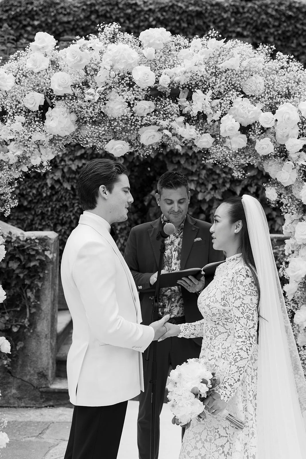 Ein Brautpaar bei einer Hochzeit, das sich die Hände hält, während der Standesbeamte im Hintergrund liest, unter einer Blumenbloc. Das Bild ist in Schwarzweiß.