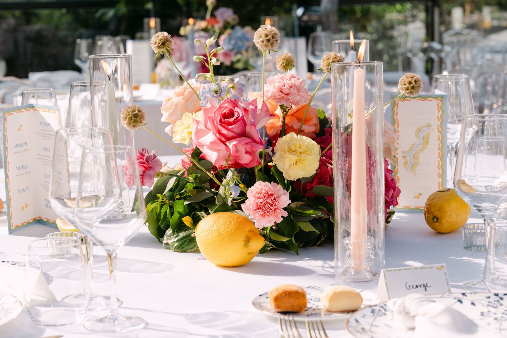 Blumenstrauß mit Rosen, Nelken und anderen Blumen auf gedecktem Tisch bei einem festlichen Anlass, umgeben von Kerzen, Weingläsern und Zitronen.