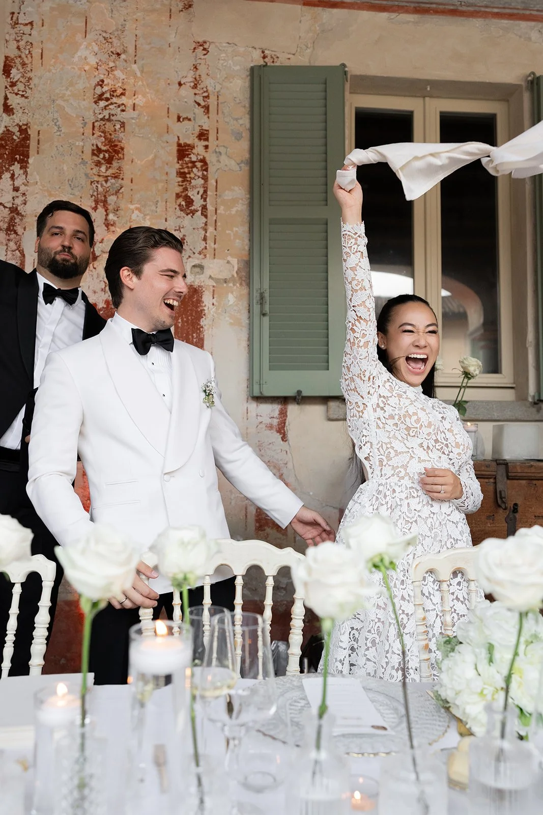 Hochzeitsszene mit lachendem Brautpaar, einer Trauzeugin, die Gegenstand in der Hand hält, und einem weiteren Mann im Anzug, im Raum mit rustikaler Wand und gedecktem Tisch mit Kerzen und weißen Rosen.