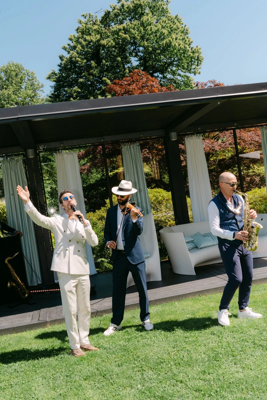 Musiker performing im Freien auf einer grünen Wiese, mit einem Mann, der singt, einen mit einer Geige und einen mit einem Saxophon. Hintergrund mit Bäumen und offenen Himmel.