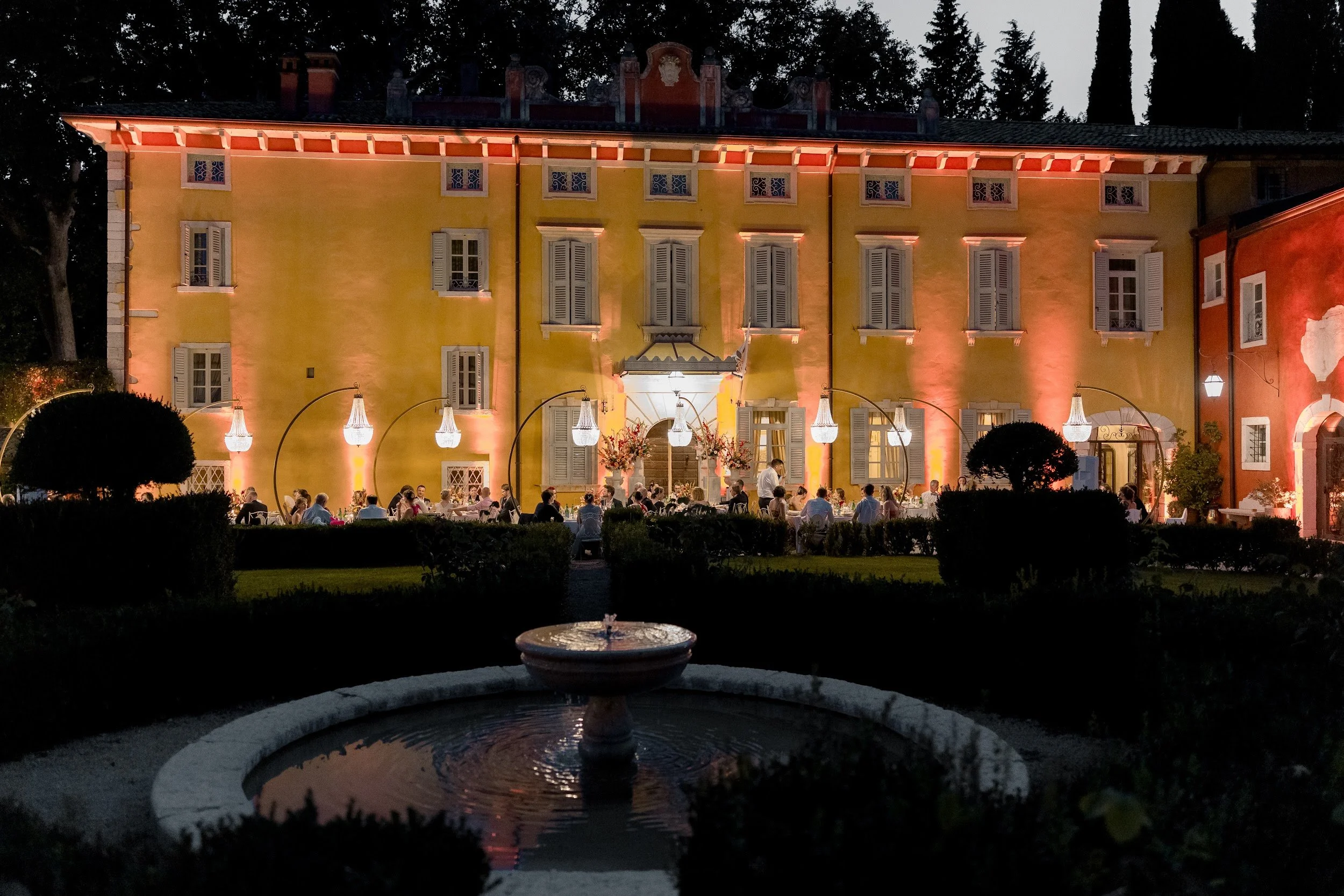 Ein beleuchtetes, gelb gestrichenes Gebäude im nächtlichen Garten mit offenstehenden Fenstern, davor eine Gruppe von Menschen, die an einem Fest oder Dinner teilnehmen. Im Vordergrund eine Brunnenanlage.