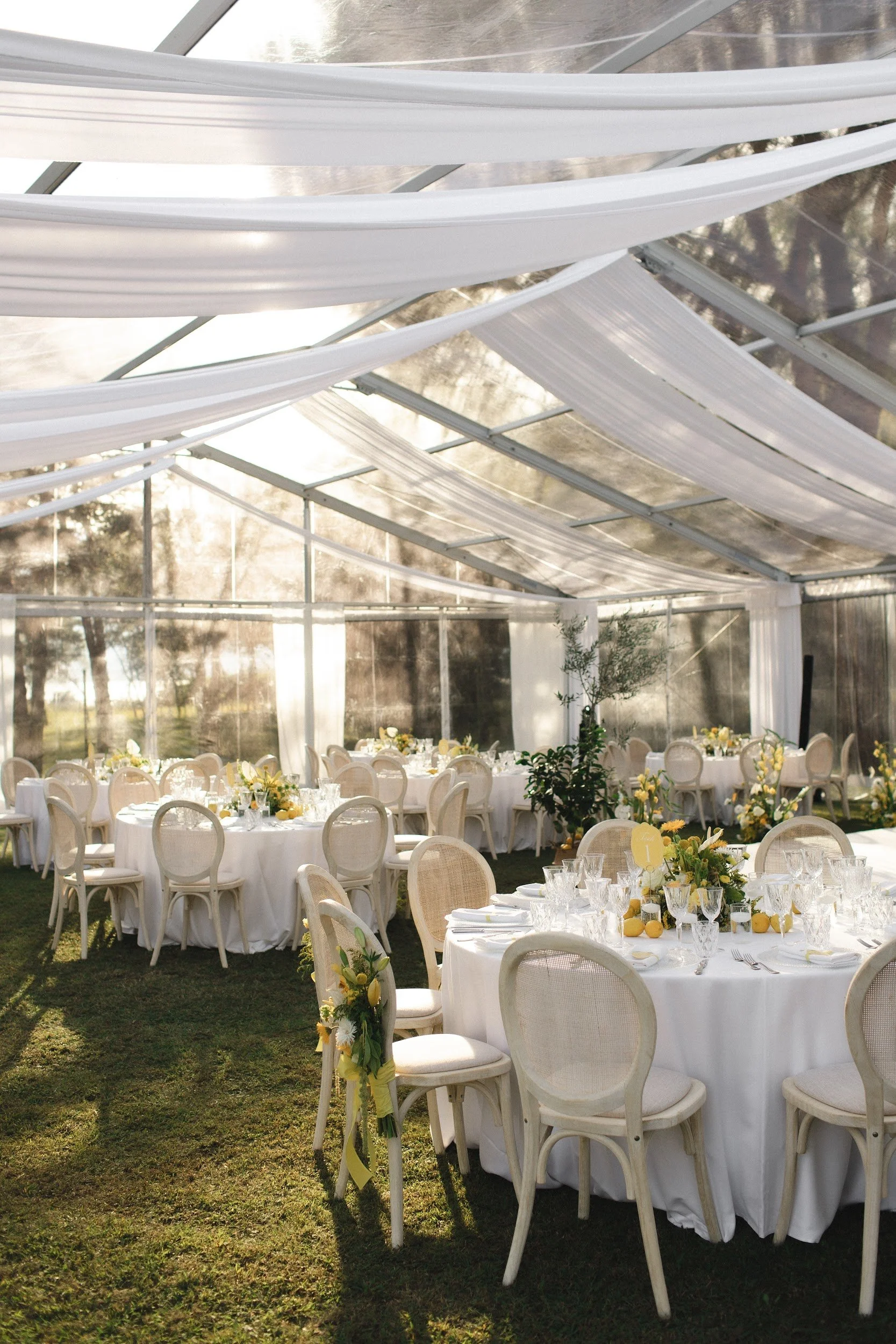 Eleganter Festsaal mit runden Tischen, weißer Tischdekoration und Blumen, in einer lichtdurchfluteten Glashalle im Freien, für eine Feier vorbereitet.