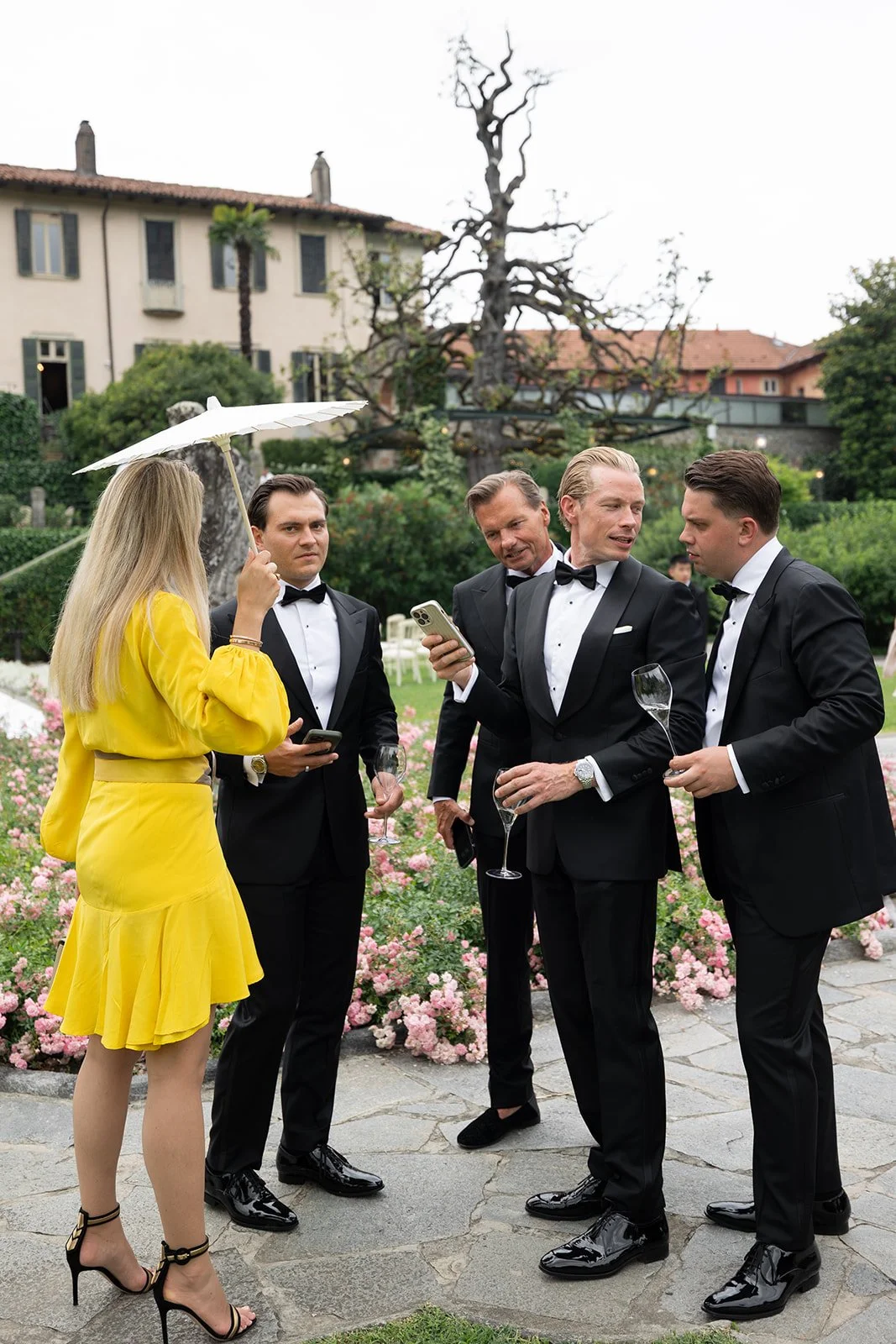 Gruppe von fünf Menschen bei einer Gartenparty, vier Männer in schwarzen Smokings mit Fliegen, eine Frau in einem gelben Kleid. Sie halten Gläser mit Sekt, sprechen miteinander, während der Mann in der Mitte sein Handy zeigt. Im Hintergrund sind Bäum