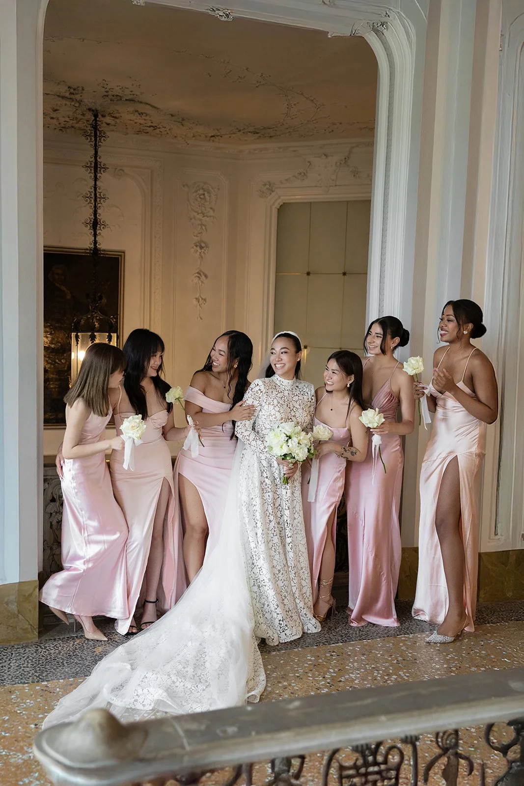 Eine Braut in einem hohen, weißen, Spitzenkleid mit langem Zug und sechs Brautjungfern in pinken, glänzenden Kleidern, die lachen und sich um die Braut versammeln, während sie in einem eleganten Raum stehen.