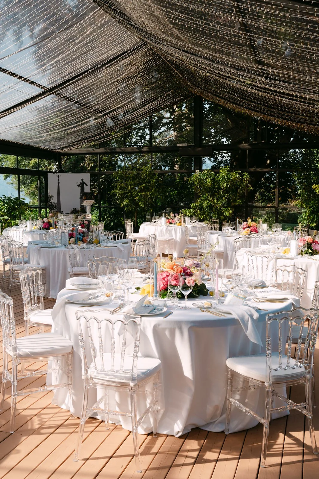 Eleganter, festlich gedeckter Veranstaltungsraum mit weißen Tischdecken, Blumenarrangements und Gläsern, umgeben von transparenten Stühlen, in einem Sonnenlicht durchfluteten Raum mit großen Fenstern und grünen Bäumen im Hintergrund.