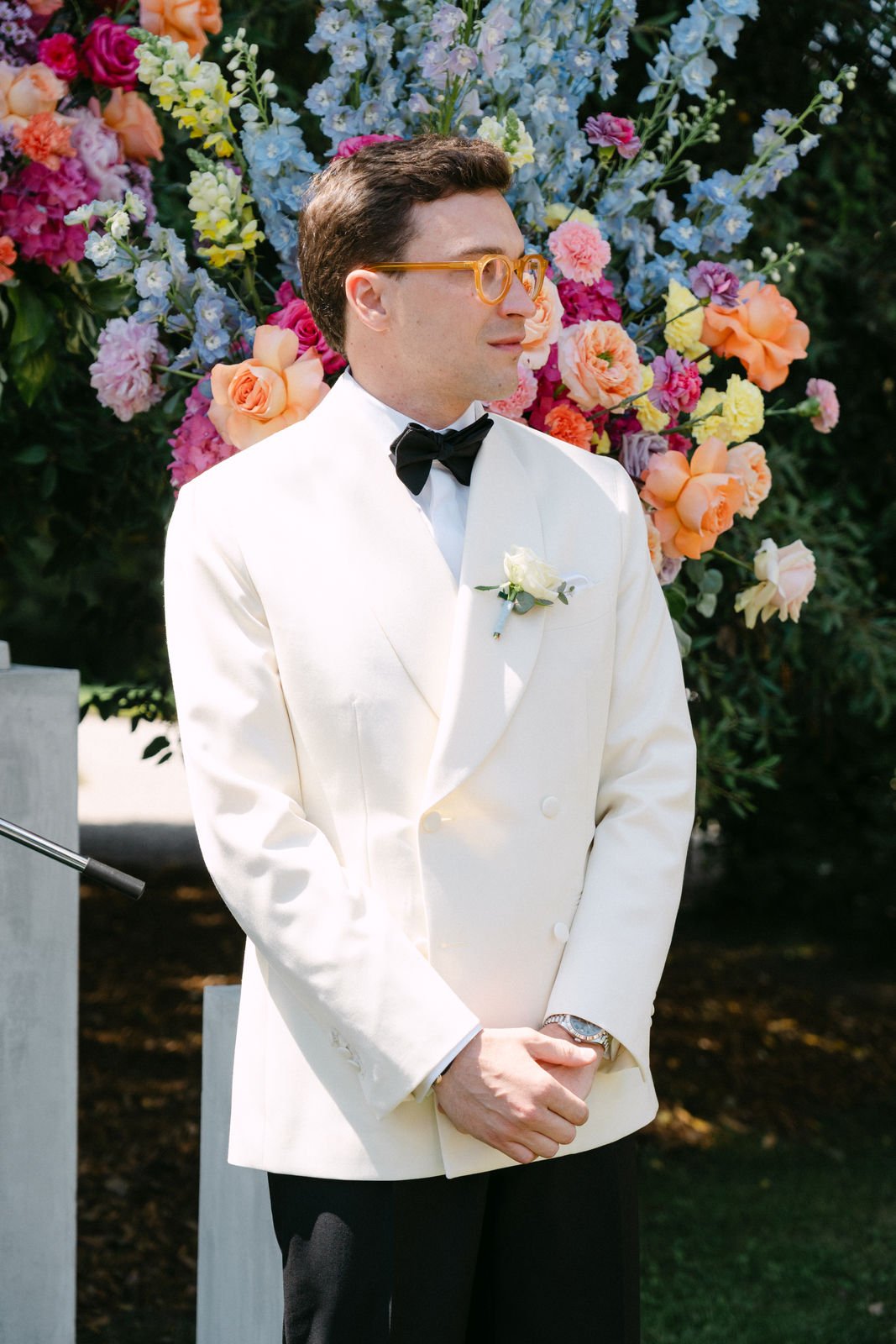 Ein Mann in formeller weißer Jacke mit Fliege steht bei einer Gartenhochzeit vor einem großen bunten Blumenarrangement.