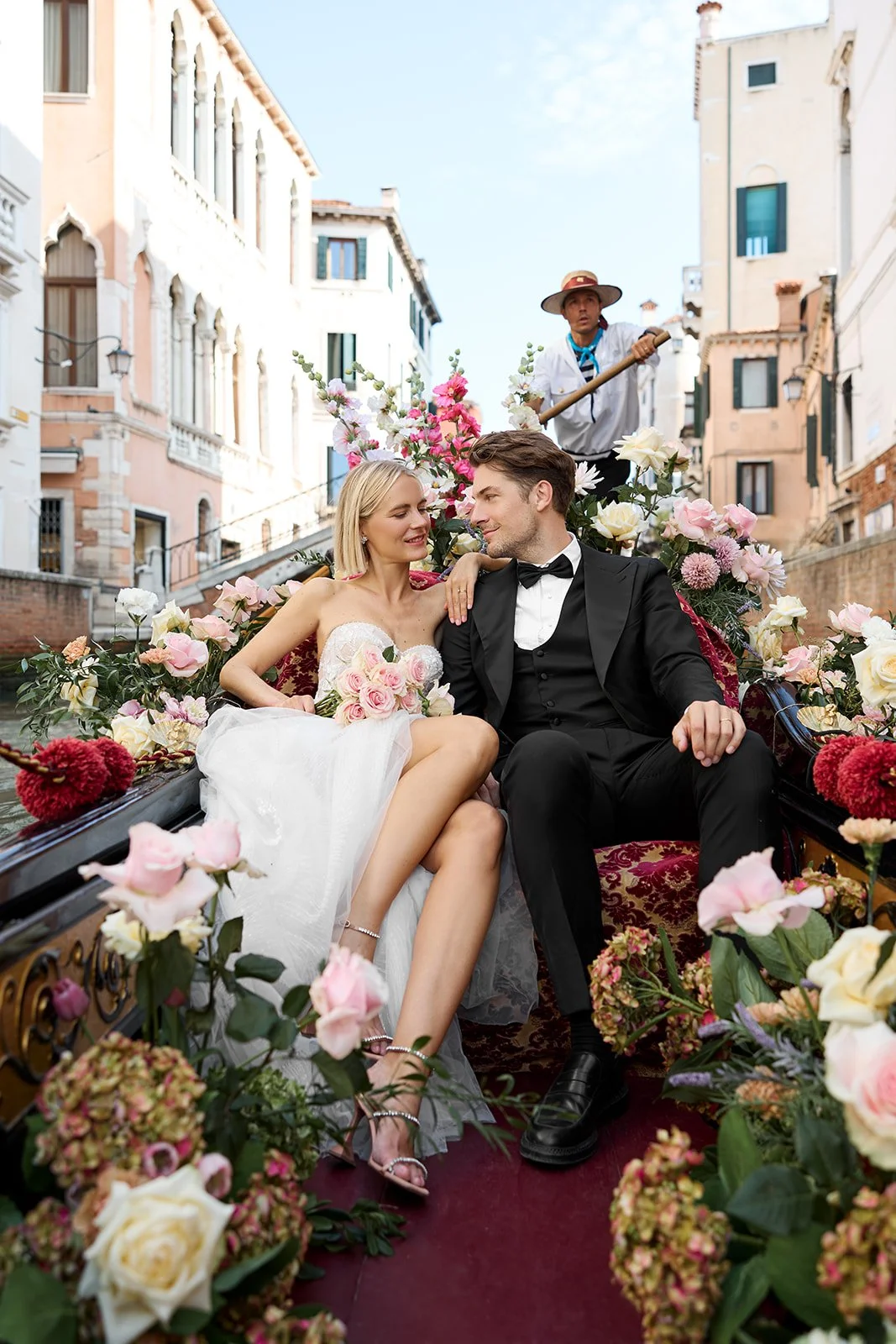 Ein glückliches Hochzeitspaar sitzt in einer Kutsche voller Blumen in einer Stadtstraße, während ein Kutscher im Hintergrund fährt.