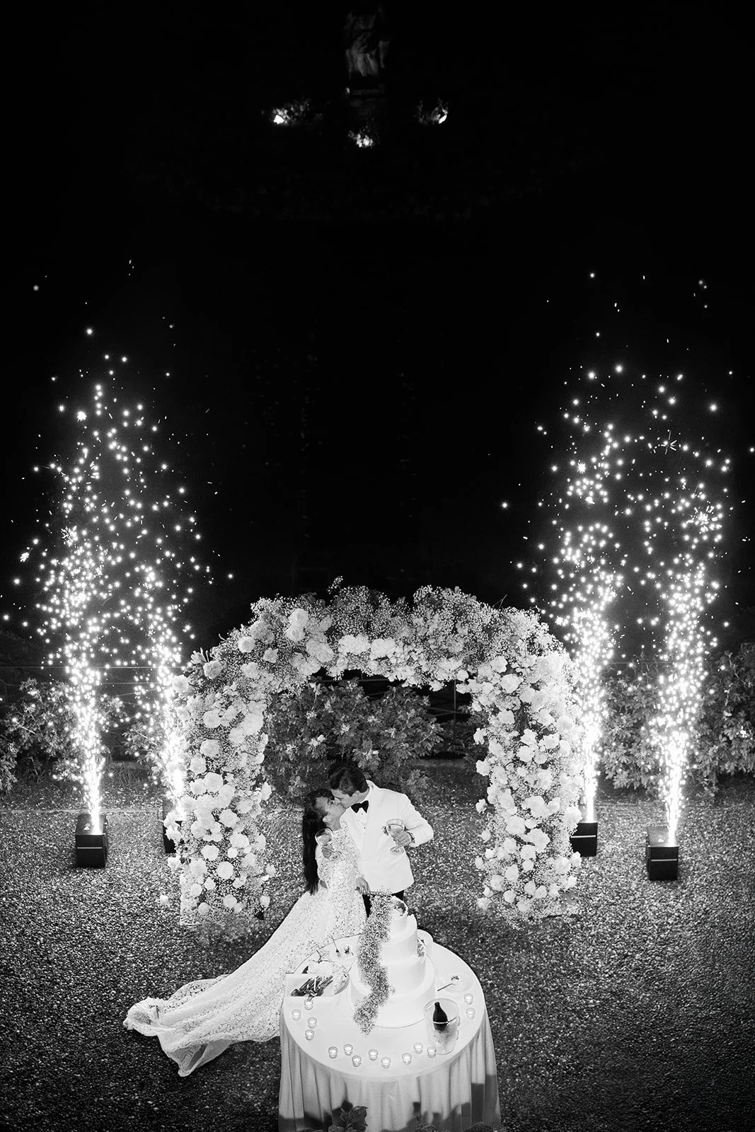 Ein Brautpaar bei der Hochzeit, das sich küsst, vor einem Blumenbogen, mit Feuerwerkskörpern im Hintergrund.