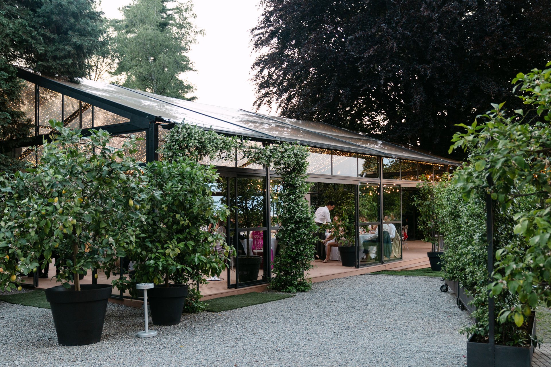 Ein Außenbereich eines Restaurants, umgeben von großen pfeffer- und zitronenbäumchen in Töpfen, mit einem gläsernen Speisesaal im Hintergrund, beleuchtet mit Lichterketten, während Menschen dort essen.