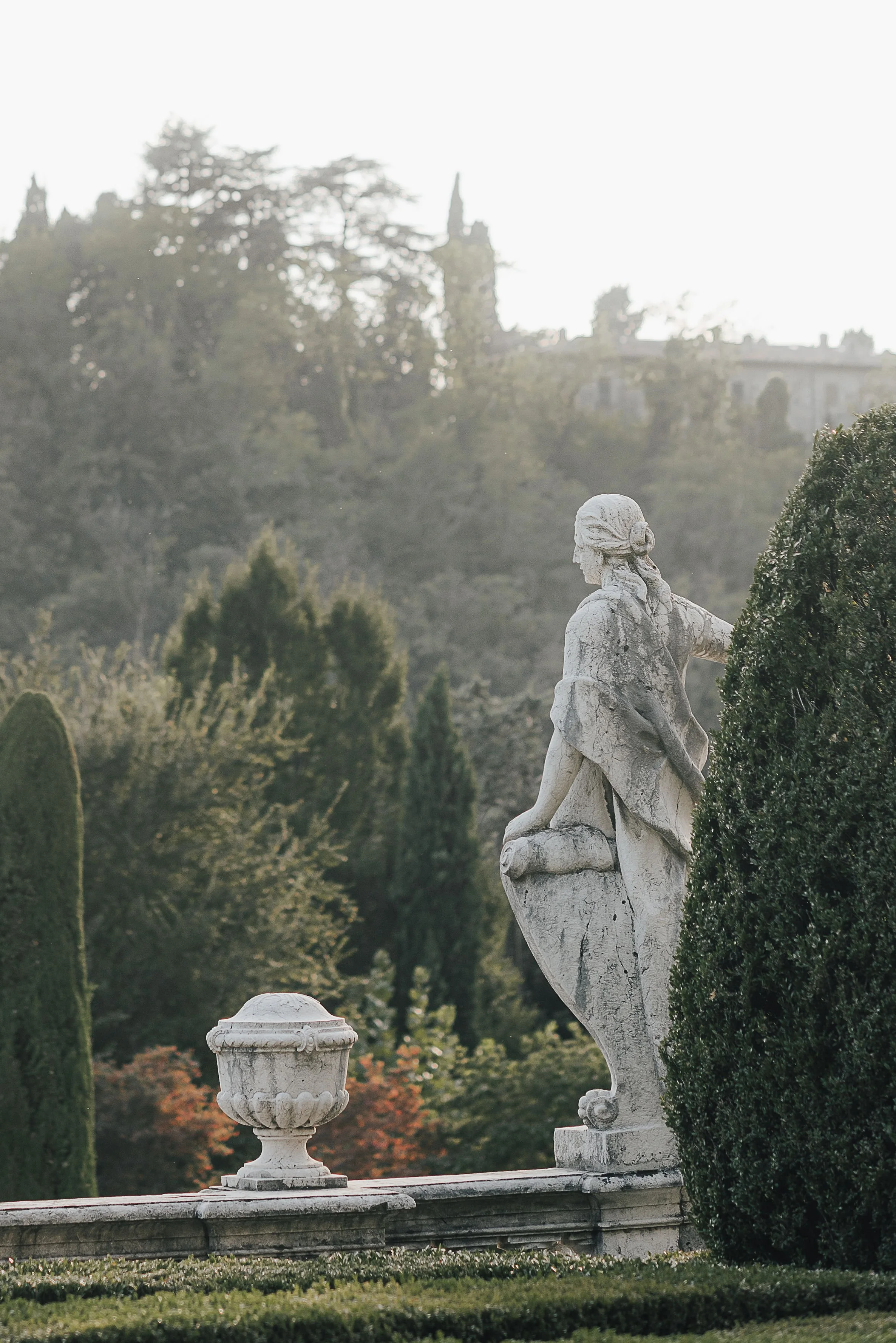 eine weiße Skulptur einer sitzenden Frau im Garten, umgeben von Bäumen und Buschwerken, im Hintergrund eine Burg auf einem Hügel