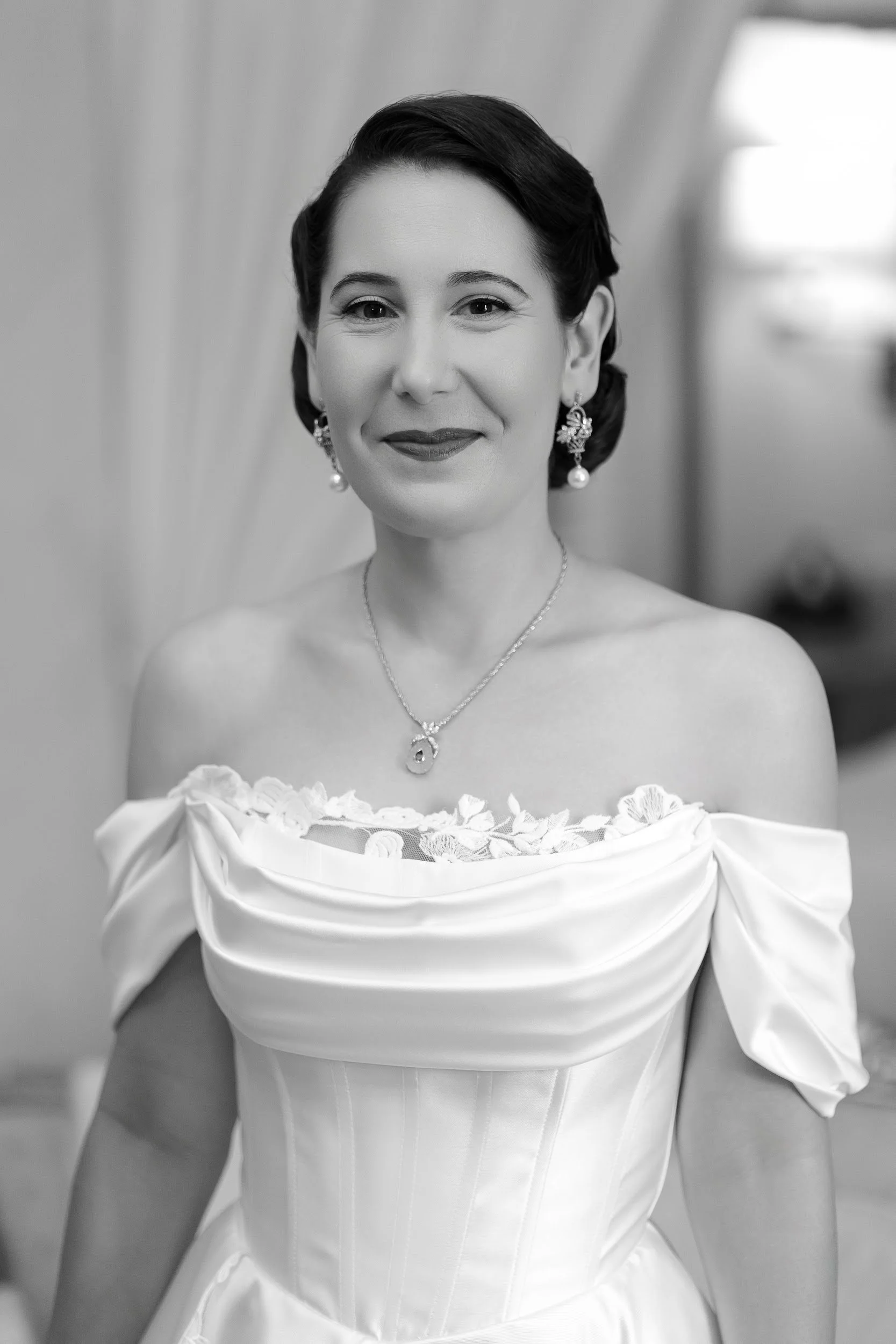 Schwarz-weiße Hochzeitsfotografie einer lächelnden braut in einem Hochzeitskleid mit Schmuck, vor einem unscharfen Hintergrund.